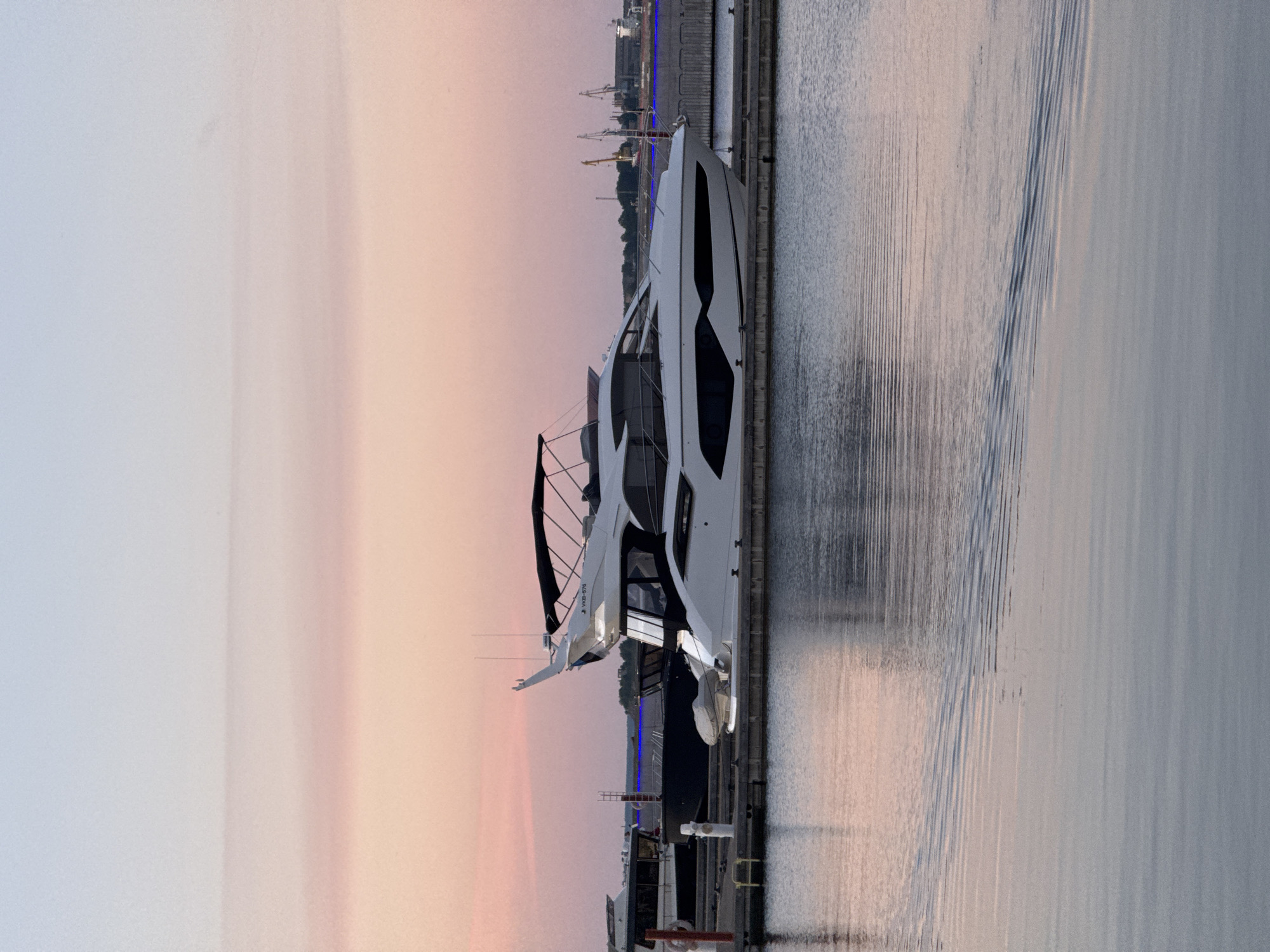 The image depicts a marina in Tallinn, Estonia, at dusk. A sleek, modern white yacht is prominently docked, reflecting in the calm water. The sky is a gradient of soft pink and blue, characteristic of a sunset or sunrise. Other boats are visible in the background, as well as industrial cranes and structures, suggesting proximity to a port or industrial area. The overall scene is peaceful and serene, with the gentle ripples on the water adding a dynamic element. There are no people visible in the image.