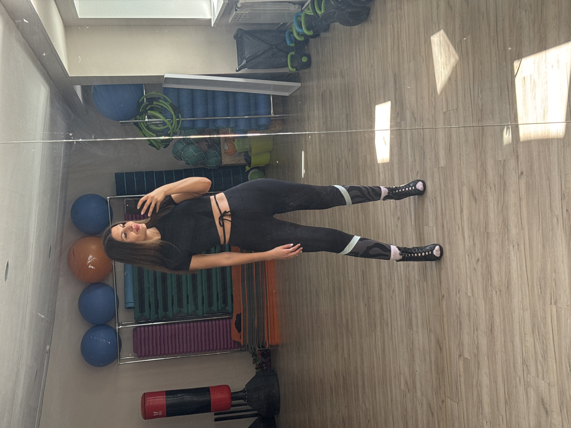 A woman is visible in a mirrored reflection in what appears to be a gym or fitness studio. She is lying on her side wearing dark workout clothes, consisting of a top and leggings with patterned accents near the ankles. Her hair is dark and straight, and she is holding a cell phone.
The background shows exercise equipment, including balls, exercise mats, step platforms, and other gym-related items arranged on shelves. The flooring is light-colored wood or laminate. Bright sunlight streams in through windows, casting sharp shadows across the floor.
The image does not provide specific information confirming the location in Chaikovsky, Russia, or details about actions, interactions, or forensic details.
