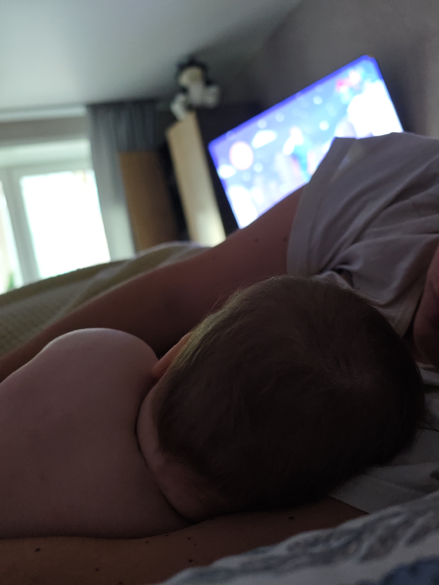 A baby lies asleep on a person's chest in a dimly lit room. A television screen illuminates the background, and a window provides natural light. A stuffed animal sits atop a shelf. The location cannot be determined from the image.