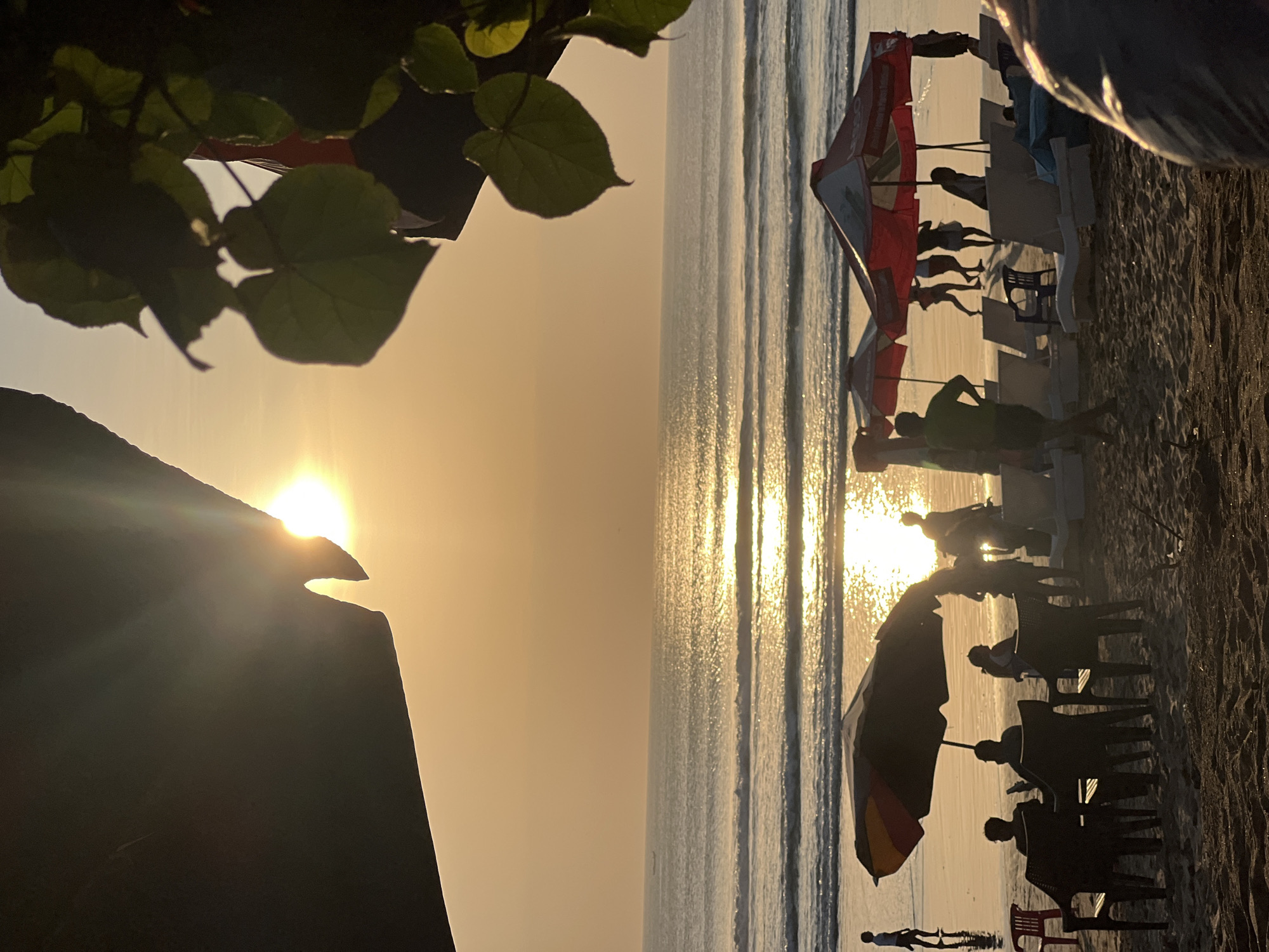 This image depicts a beach scene in Kuta, Indonesia, at sunset. The sun, a bright orb, is low on the horizon, casting a golden glow across the sky and water. Gentle waves roll onto the sandy shore. In the foreground, several beach umbrellas and chairs are set up. Silhouetted figures of people are visible, some seated at tables under umbrellas, others walking or standing near the water's edge. Lush green leaves from an overhanging tree frame the top right portion of the image. The overall atmosphere is tranquil, with the warm hues of the sunset creating a peaceful ambiance.