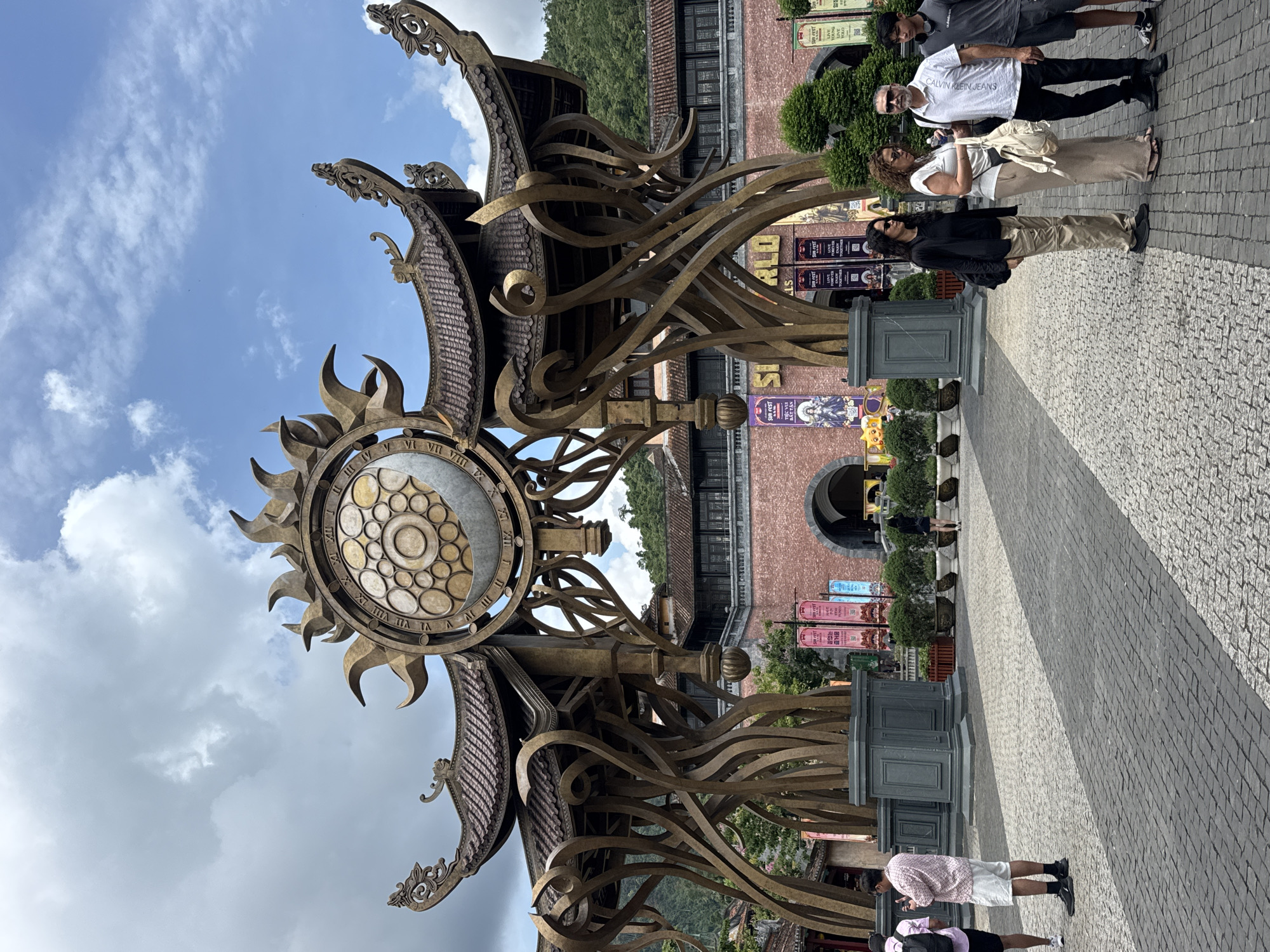 The image shows a large, decorative sundial-like structure in Đà Nẵng, Vietnam. The structure features a clock face with Roman numerals surrounded by sun-like rays, set atop a Vietnamese-style roof and wave-like metal supports. A brick building with arched entrances is visible in the background, with banners hanging in front. Several people are present: one group of four standing on the right side of the frame, and another smaller group below and to the left. The ground is paved with interlocking stones. The sky is partly cloudy.