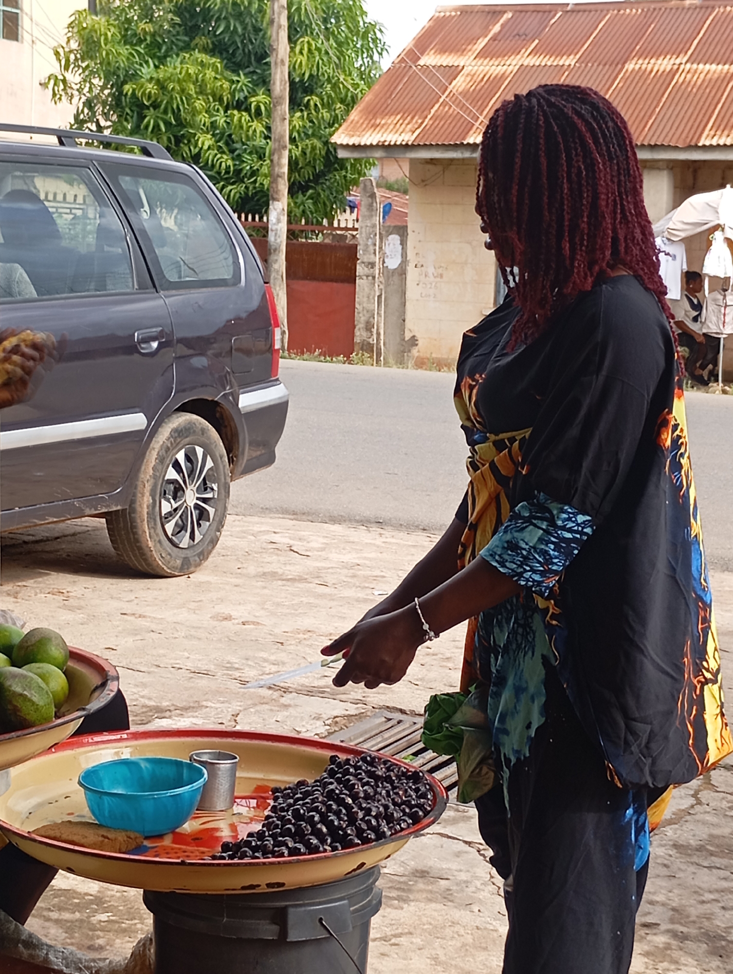 This media file shows a side view of a Black woman with dark red, braided hair, wearing a colorful, patterned garment. She appears to be a vendor, standing behind a display of goods. In the foreground, a round metal tray sits on top of a black bucket. The tray holds a blue bowl, a small metal cup, and a pile of dark, round berries. To the left of this tray is another similar tray holding several green fruits, possibly avocados or mangoes. The woman is holding a thin, white object, possibly a receipt or a money receipt, and her right hand is reaching towards the berries.
Behind the woman and her stall, a dark gray station wagon is parked. The background reveals a street with some buildings, one with a corrugated iron roof that appears rusted. Trees with lush green foliage are visible, suggesting a tropical or sub-tropical environment. The ground is a mixture of concrete and sand. The lighting suggests it is daytime, possibly mid-morning or afternoon, with overcast skies or diffused sunlight. The overall scene appears to be a street market or roadside stall in a location like Jos, Nigeria. There is no visible text in the image.