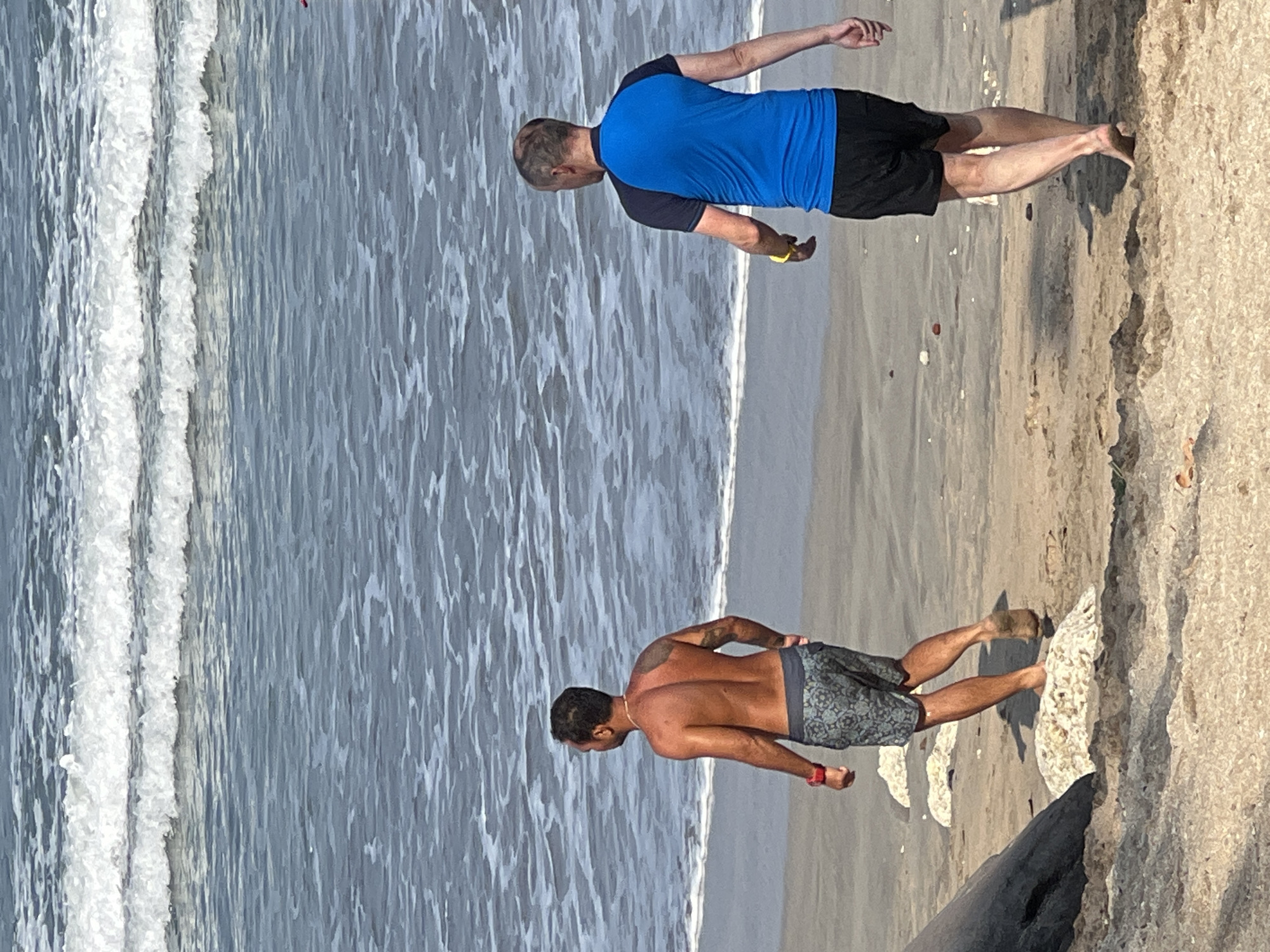 Two men are walking on a sandy beach. The man on the left is shirtless and wearing patterned swim shorts. He has a watch on his left wrist. The man on the right is wearing a blue and black rash guard and black swim shorts. He has a yellow band on his left wrist. The ocean is behind them with waves breaking on the shore. The sand is light brown and there are some scattered rocks and debris. The image appears to be taken on a sunny day. The location is the city of Kuta, Indonesia.