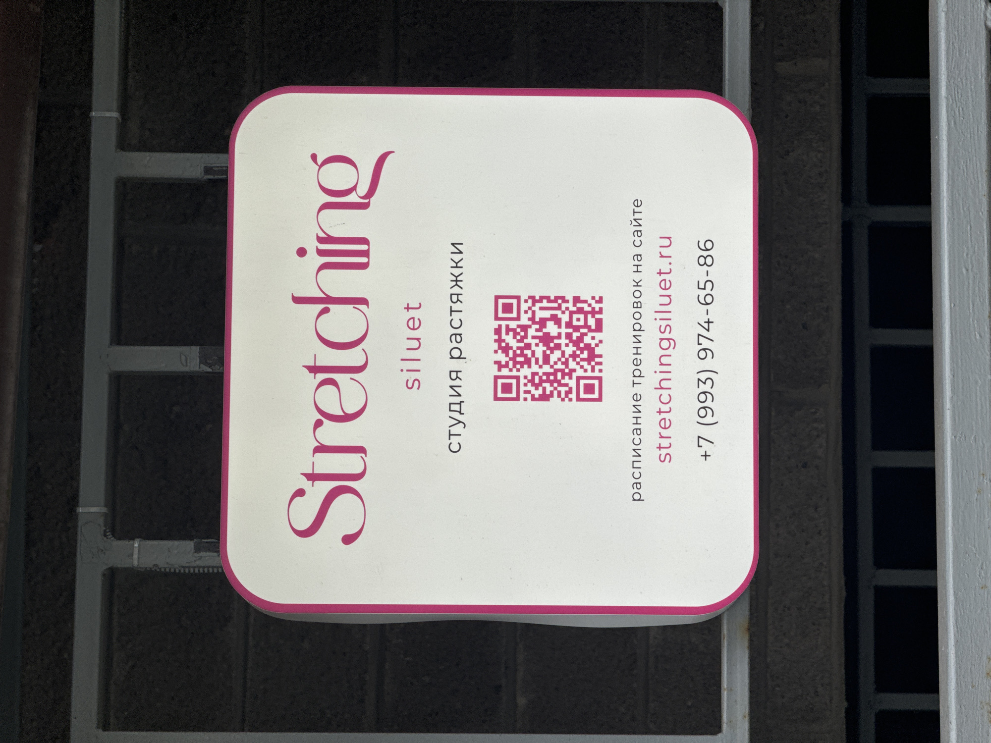 The image displays a white, rectangular sign with rounded corners, advertising a stretching studio. The sign is positioned vertically, with its longer dimension angled from the bottom left to the upper right of the frame. The background is a dark, grid-like metal structure, possibly a fence or gate, suggesting an outdoor or semi-outdoor location.
The most prominent text on the sign is "Stretching" in a large, stylized pink font. Below this, in smaller pink text, are the words "siluet" and "студия растяжки," which translates to "stretching studio." A pink QR code is centrally located on the right side of the sign. Further right, black text reads "расписание тренировок на сайте" (training schedule on the website), followed by the website address "stretchingsiluet.ru" in pink. At the bottom right, a phone number, "+7 (993) 974-65-86," is also displayed in pink.
No people are visible. The setting appears to be urban, likely an entrance or a public space in Saint Petersburg, Russia, given the Russian text. The lighting is even, suggesting either an overcast day or the sign is in a shaded area, with no strong shadows to indicate the time of day. The sign functions as an advertisement, providing essential information for potential clients of the stretching studio.
