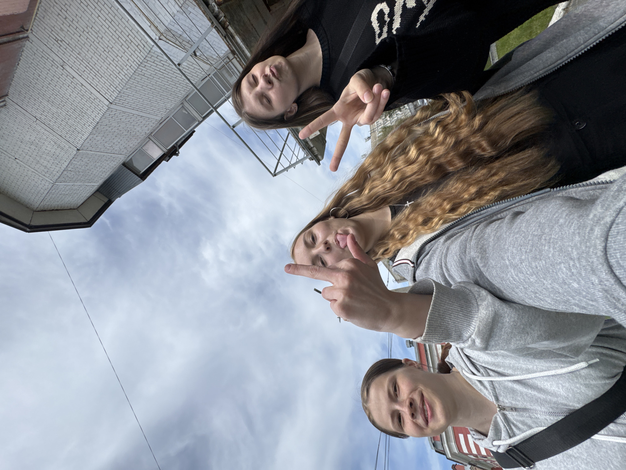 The media file is a low-angle selfie featuring three young women, with the photograph rotated approximately 90 degrees counter-clockwise. The sky, filled with scattered white clouds, forms a significant portion of the background.
In the foreground, positioned towards the bottom of the rotated image, a young woman with short, dark brown hair tied back smiles directly at the camera. She is wearing a grey hooded sweatshirt and a black strap crosses her chest.
Behind her, another young woman with long, wavy light brown hair is visible. She wears a grey hooded sweatshirt over a black top and is captured mid-gesture, sticking out her tongue and pointing her index finger upwards. A visible hoop earring can be seen.
Further back, towards the top left of the rotated image, the third young woman has long, straight dark brown hair. She is wearing a black sweater adorned with a white, stylized text or pattern on the front. Her expression is playful, with puckered lips, and she holds up her hand to make a peace sign.
The background shows elements of an urban environment. A multi-story white brick building with windows and metal support structures is prominent on the left side of the rotated frame. Hints of a red and white building are visible towards the bottom right. A thin power line or cable stretches across the cloudy sky. The overall mood is casual and friendly, characteristic of a group selfie. The time of day is daytime, under cloudy conditions.