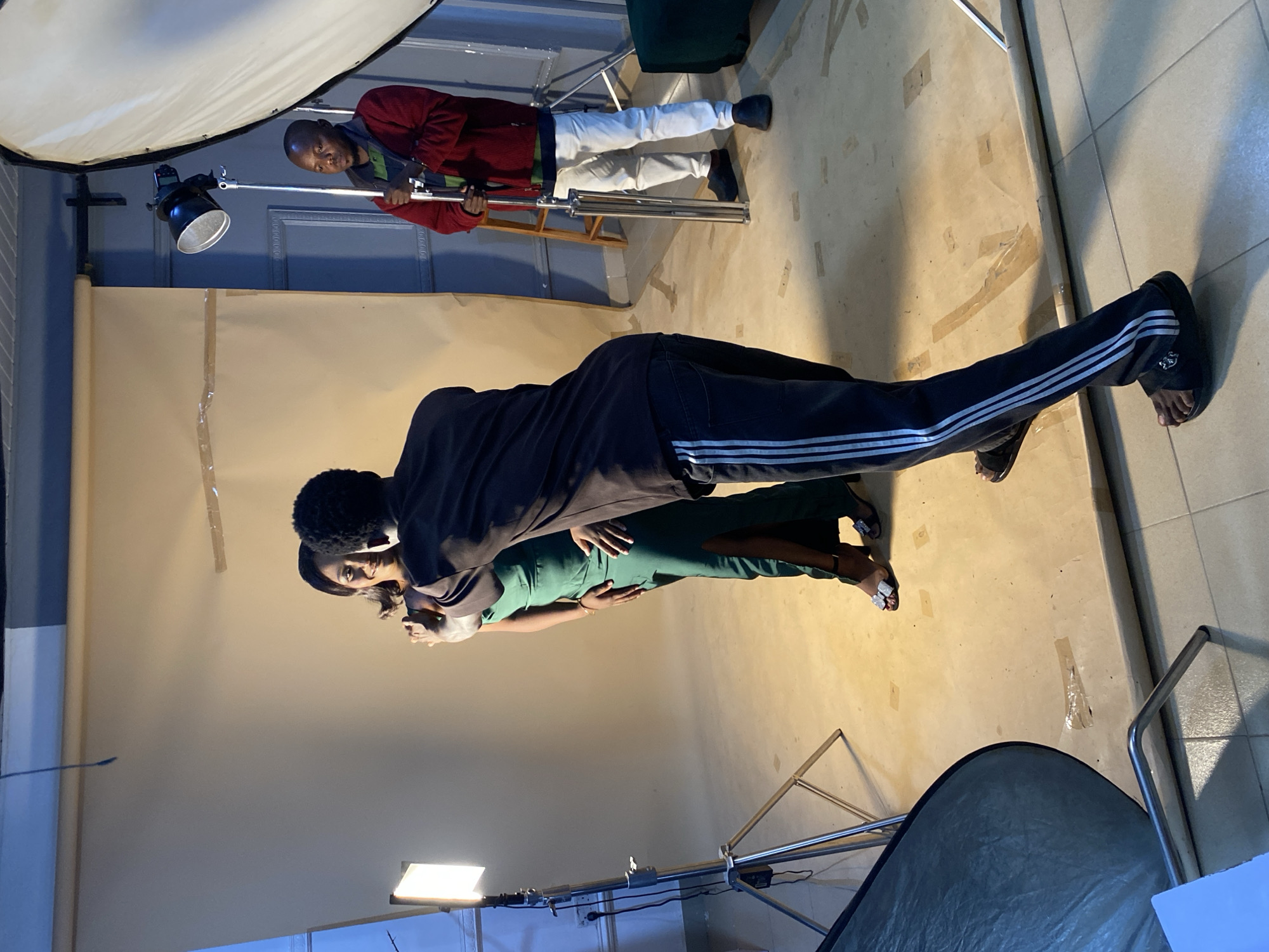 This media file captures a behind-the-scenes look at a photoshoot in a studio setting in Bukuru, Nigeria. The scene features three individuals. In the background, a man wearing a red sweater and white pants is adjusting lighting equipment, specifically a large softbox. In the foreground, a couple is posing for the camera. The man is wearing dark track pants with white stripes and a dark top, while the woman is in a fitted green dress. They are positioned closely together, with the woman touching the man's chest.
The studio appears to be a simple space with a backdrop and various professional photography lights and stands. The floor is made of tiled material. The lighting suggests an indoor, controlled environment, typical for a studio. The overall mood appears professional and focused on achieving the desired shot. There is no visible text within the image. The time of day and weather are not determinable from the image content.