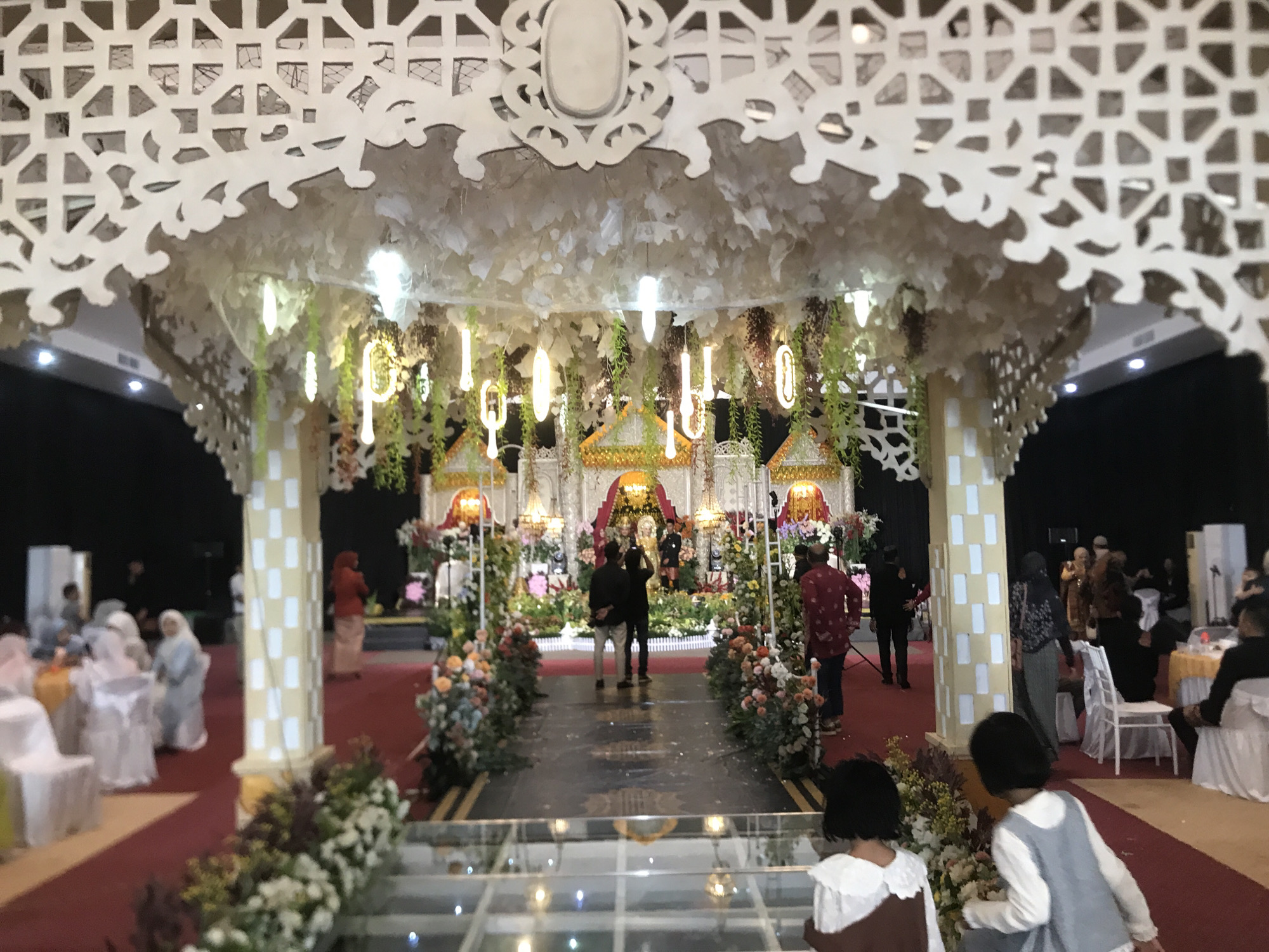 The image shows a wedding reception in Lhokseumawe, Indonesia. A decorative archway with white latticework and faux foliage frames the view of a wedding stage. Illuminated letter-shaped decorations hang above the stage. A red carpet leads to the stage, flanked by floral arrangements. Several people stand on the stage, including a group taking photos. Guests are seated at tables to the sides of the carpet. Two young girls stand in the foreground, their backs to the camera. The floor appears to be partially made of clear glass panels with decorative lighting beneath.