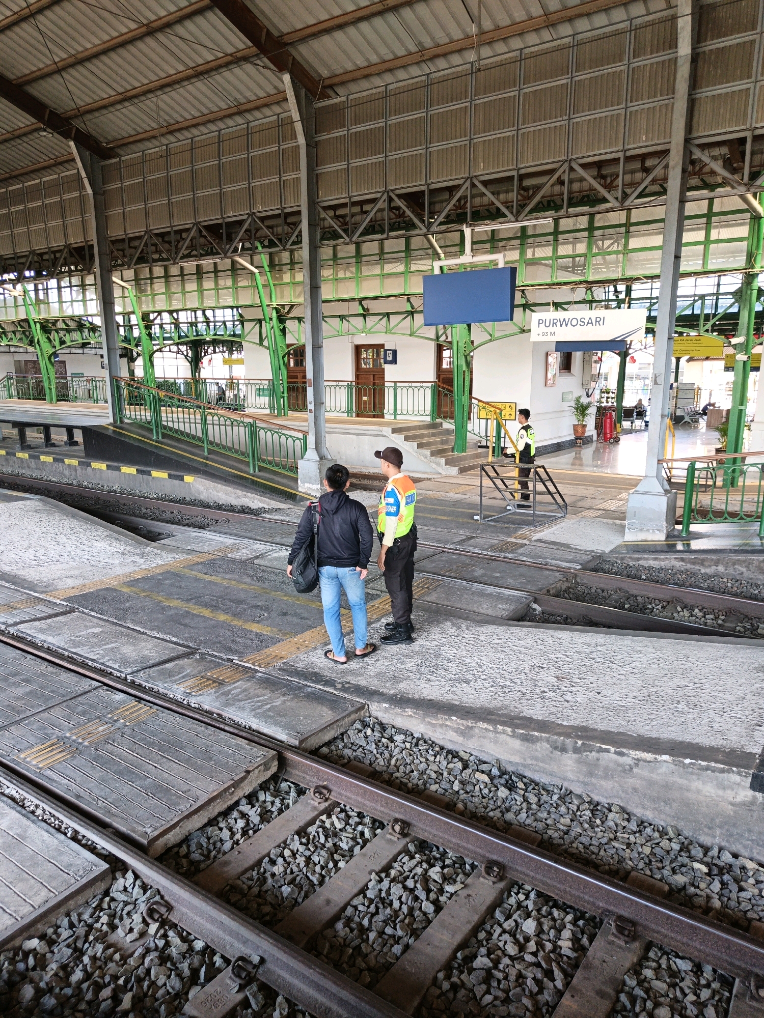 The image depicts the Purwokerto train station platform in Surakarta, Indonesia. Two individuals are standing on the platform near the train tracks. The person on the left is wearing a black hoodie, blue jeans, and flip-flops, carrying a dark bag over their shoulder. The person on the right is wearing a high-visibility yellow vest over a dark uniform and a cap, suggesting they are a railway employee. They appear to be facing each other, possibly in conversation. The platform is made of concrete with yellow tactile paving. Train tracks with gravel ballast are visible in the foreground and between the platform. The station has a covered roof supported by green and grey metal structures. A sign in the background reads "PURWOSARI". Other elements visible include railings, stairs, a small building with doors, and some seating areas in the distance. The overall environment is a functional train station platform on a bright day.