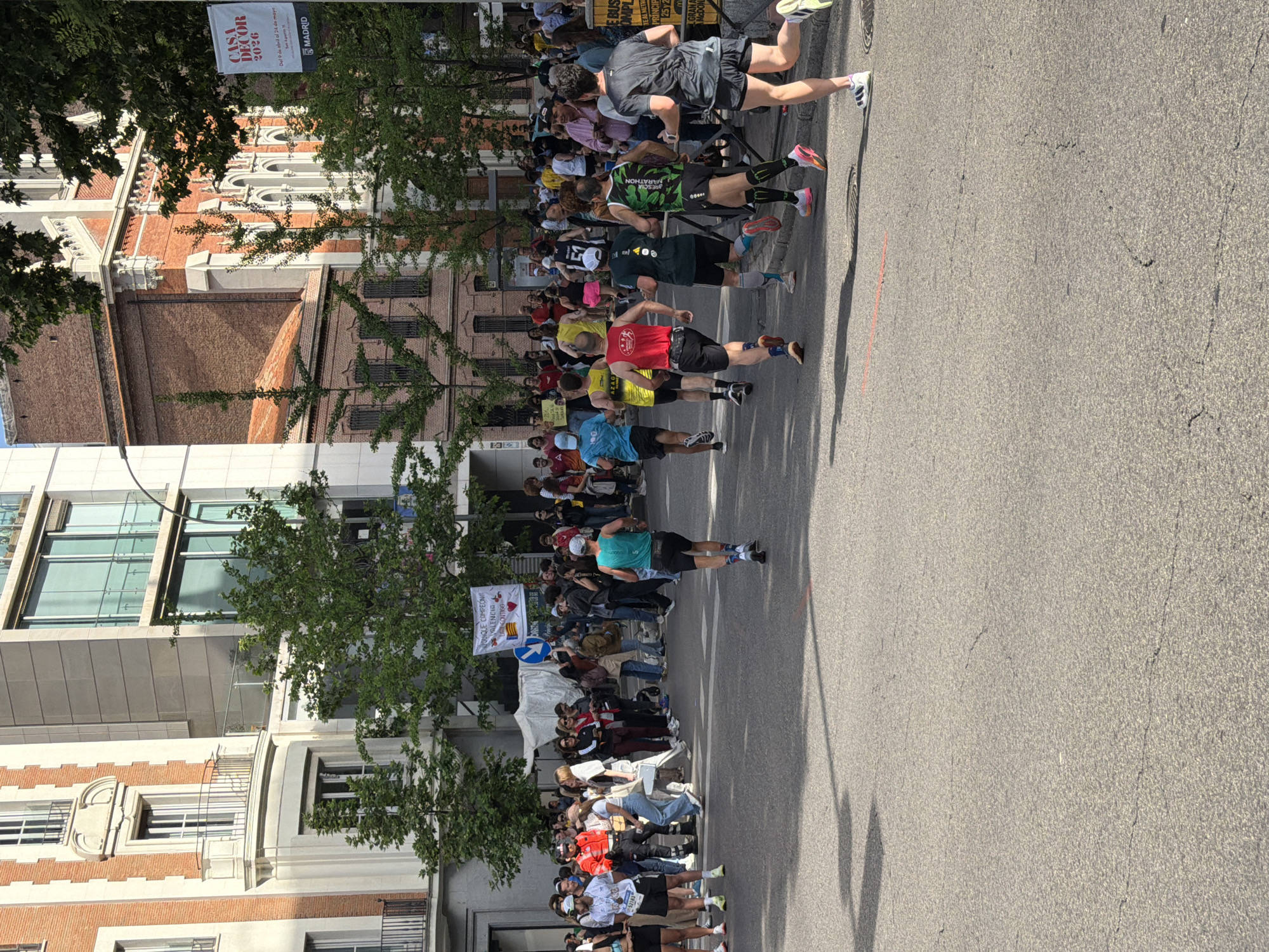 Here's a detailed description of the image:
**Scene:** The image captures a street scene, likely a marathon or road race, taking place in the city of Madrid, Spain. The road is wide and asphalted, running parallel to the buildings.
**People:** A large crowd of runners is visible. They are wearing various colored athletic clothing. Spectators are present on the sidewalks, lining the route to watch and cheer.
**Buildings:** A mix of architecture styles is visible, including older brick buildings, with arched windows, alongside more modern structures. One building appears to have a modern glass facade. Banners related to "CASA DECOR 2026" and Madrid are visible.
**Objects:** Trees line the street on both sides providing shade. There is a sign held by spectators supporting "QUIQUE CAMPEON," with "VALENCIA" written on it. There is also a bus nearby.
**Actions:** The runners are in motion, running along the road. Spectators are watching and cheering them on.
**Environment:** The scene is brightly lit suggesting it is daytime.