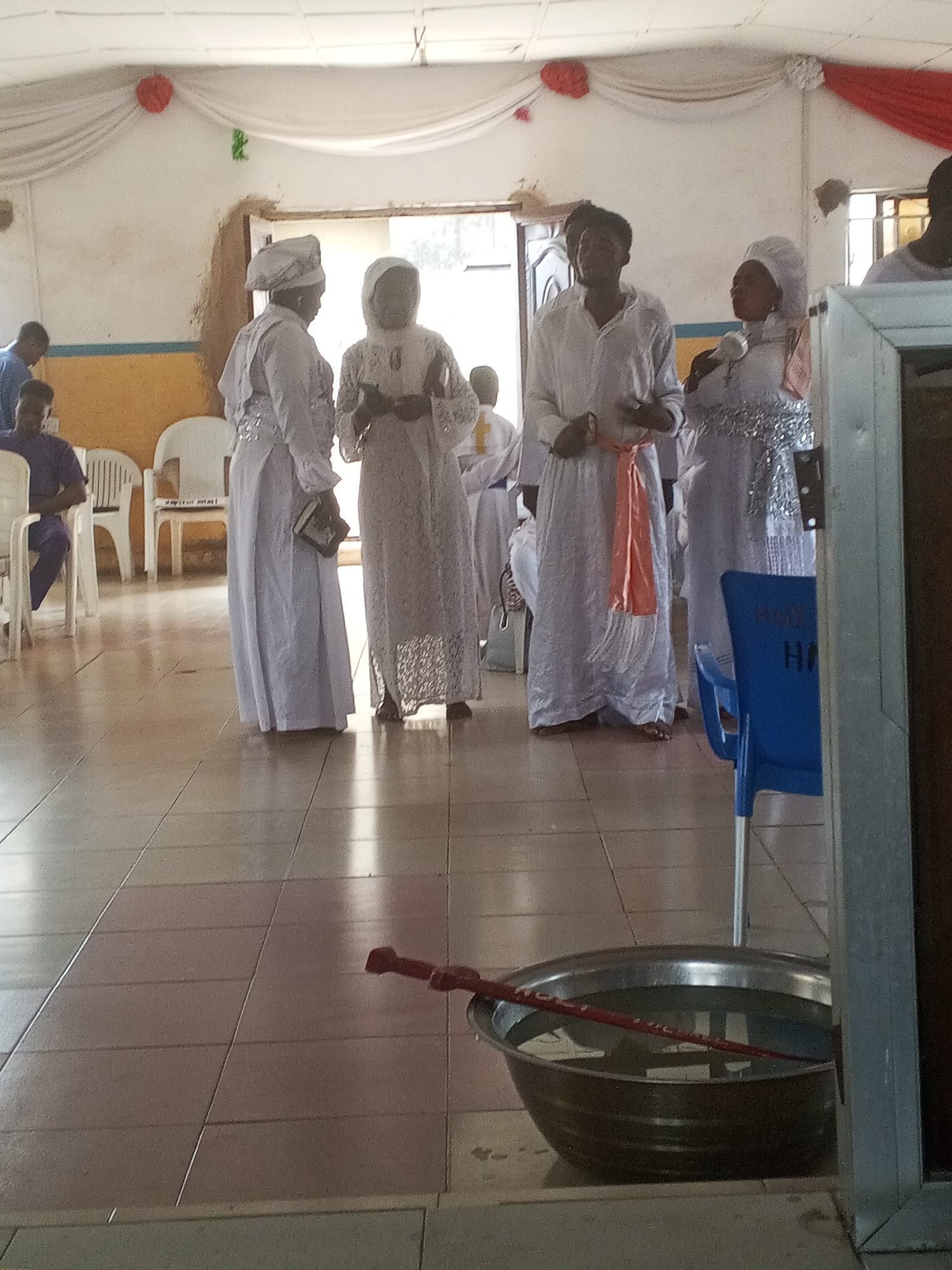 The image shows an indoor scene in what appears to be a church or religious gathering in Lokoja, Nigeria. Several people are present, most of whom are wearing long, white robes and head coverings. A man in the center is wearing a white robe with an orange sash. They are standing near a doorway or opening. In the foreground, there is a metal bowl filled with water and a red object submerged within it. The floor is tiled. White fabric drapes decorate the upper part of the wall, with some red decorations. Several chairs are visible in the background.