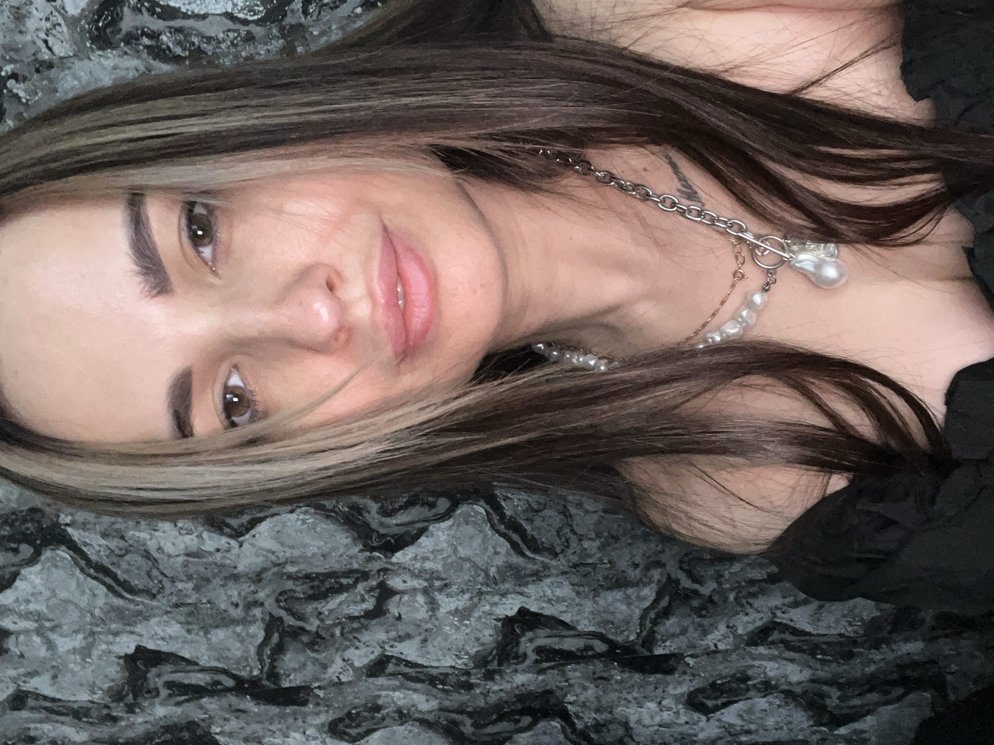 A woman with brown hair and light brown highlights is lying on a surface with a gray marble-like pattern. She is wearing two necklaces: one with a silver chain and a large pearl pendant, and another with smaller pearls. She has brown eyes, and her lips are slightly parted. The background is out of focus. The location is Unknown, Unknown.
