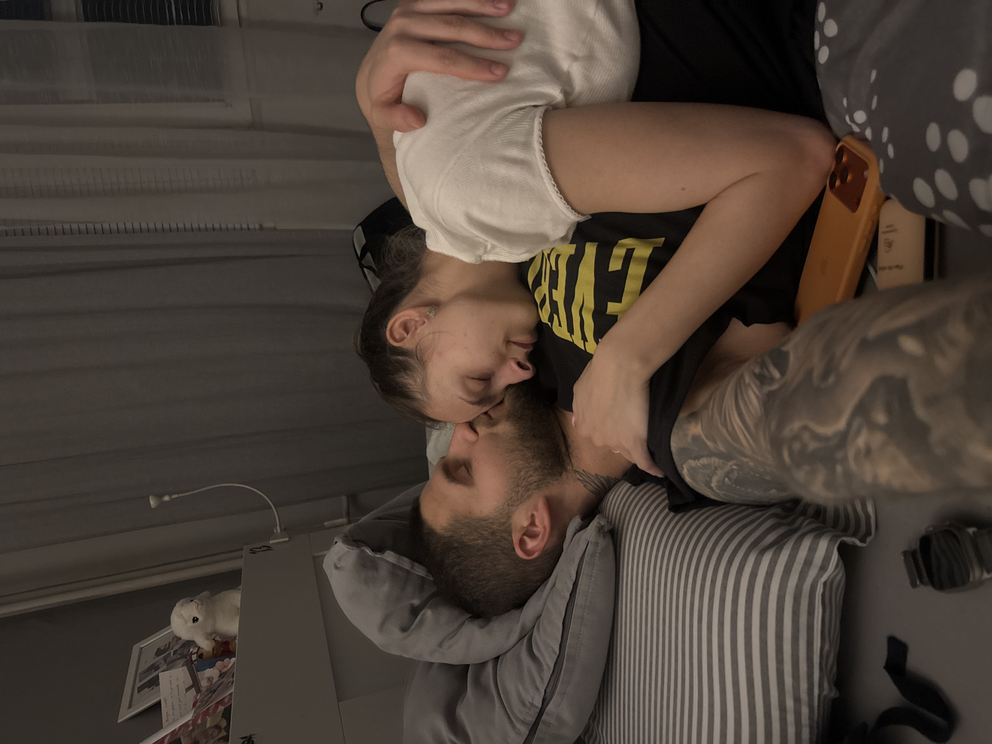 A man and a woman are lying in bed, facing each other, with their faces close together in an intimate moment. The man is lying on his back, with his head resting on a gray pillow with white stripes. He has short, dark hair and a beard. The woman is lying on top of him, partially covering his face. She has dark hair pulled back and is wearing a white shirt. The man's arm is visible with a detailed tattoo.
On the bed, there are different patterned pillows. To the left, a gray shelf holds a lamp, a stuffed animal, and a framed picture. The scene is indoors, likely in a bedroom. There is no direct indication that the scene is specifically in Sofia, Bulgaria.