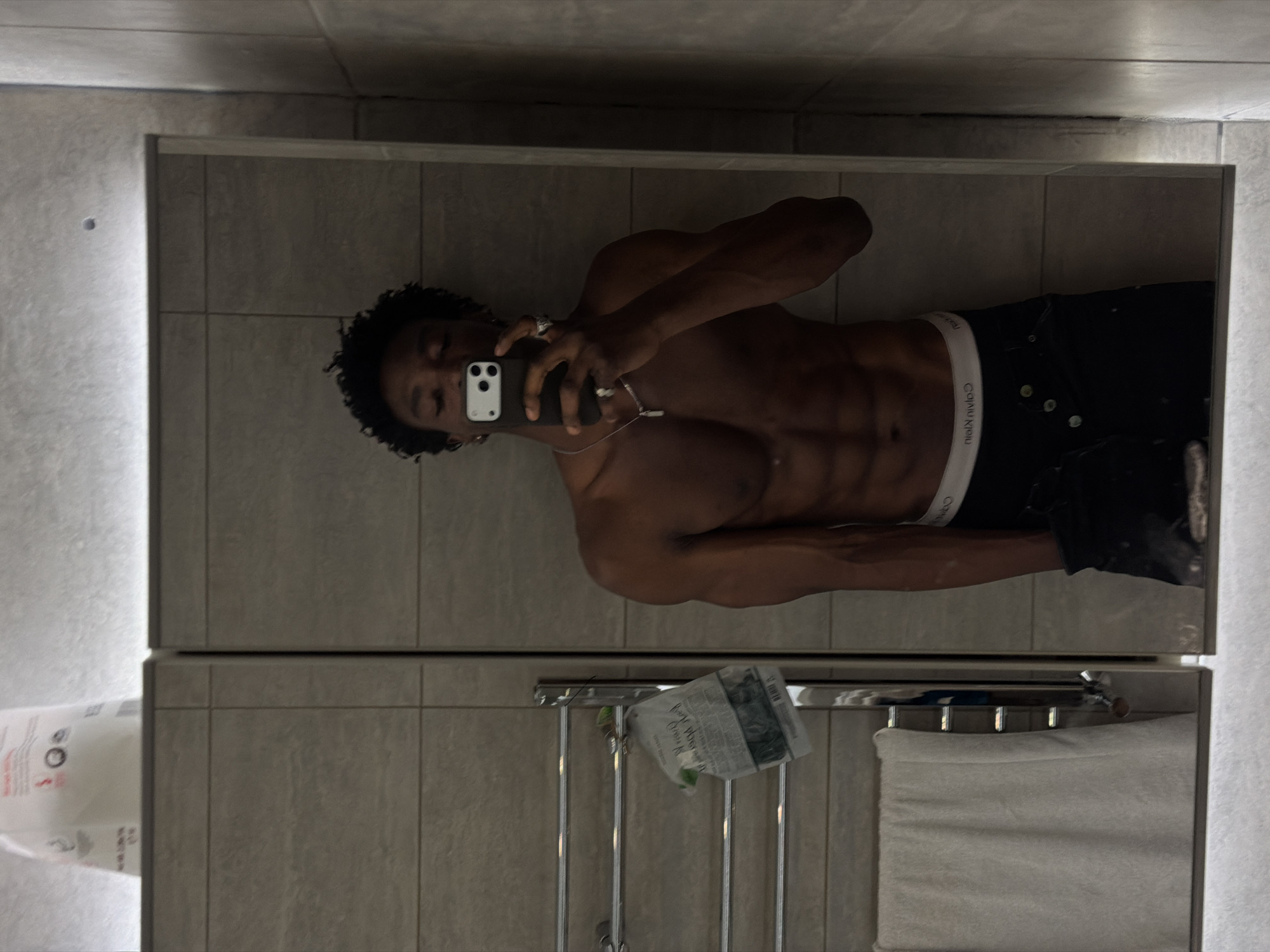 A shirtless man with dark curly hair takes a mirror selfie in a bathroom. He is wearing black jeans with a white Calvin Klein waistband. A towel rack holds a newspaper and a white towel. The bathroom is tiled in grey. The location is indeterminable from the available visual information.