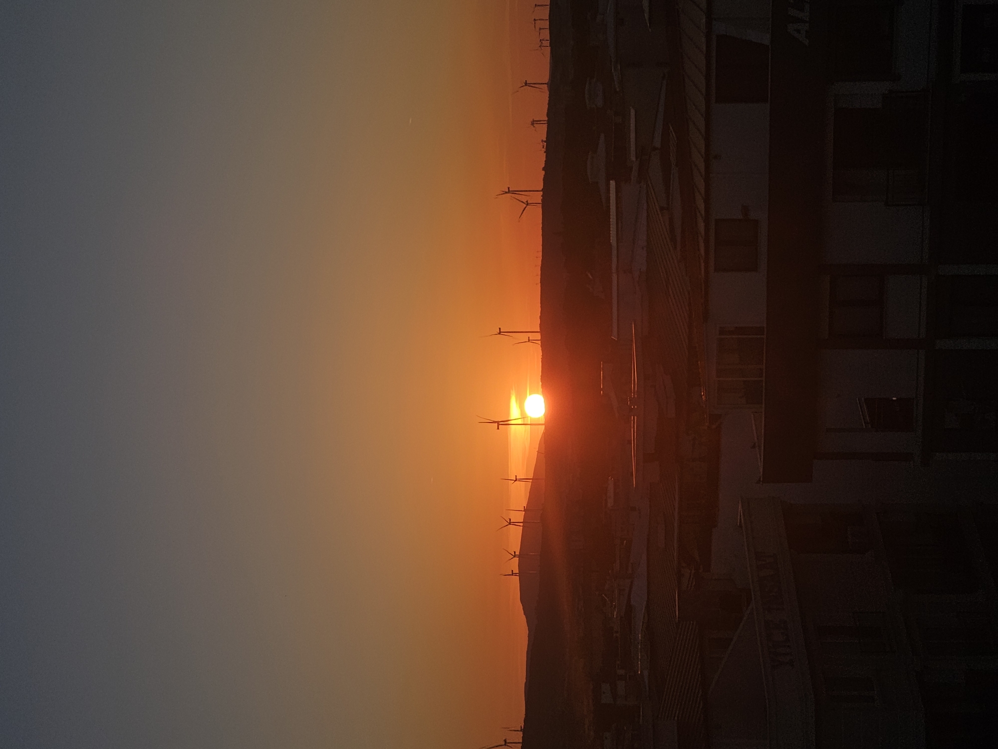 This image captures a serene sunset or sunrise over Seferihisar, Turkey, presented in a landscape orientation (rotated from the original upload). The sky is dominated by a vibrant gradient, transitioning from a deep orange and yellow near the horizon, where the sun is visible, to a softer grey-blue at the top of the frame.
The sun, a brilliant, almost white orb, is positioned low on the horizon, partially obscured by a dark, silhouetted mountain range or ridge line that stretches horizontally across the midground. Along this ridge, numerous wind turbines stand as dark, distinct silhouettes against the brightly lit sky, emphasizing renewable energy infrastructure within the natural landscape.
In the right foreground, several large, dark buildings are silhouetted, their shapes sharply defined against the glowing sky. These structures appear to be industrial or commercial, with visible windows and structural details hinted at by the faint light. Partial, obscured text, including the letters "AL", can be discerned on the side of one of these buildings.
No people are visible, and no specific activity is taking place, contributing to the tranquil atmosphere. The weather appears clear and calm. The scene combines natural beauty with human-made structures, capturing the moment of transition between day and night.
