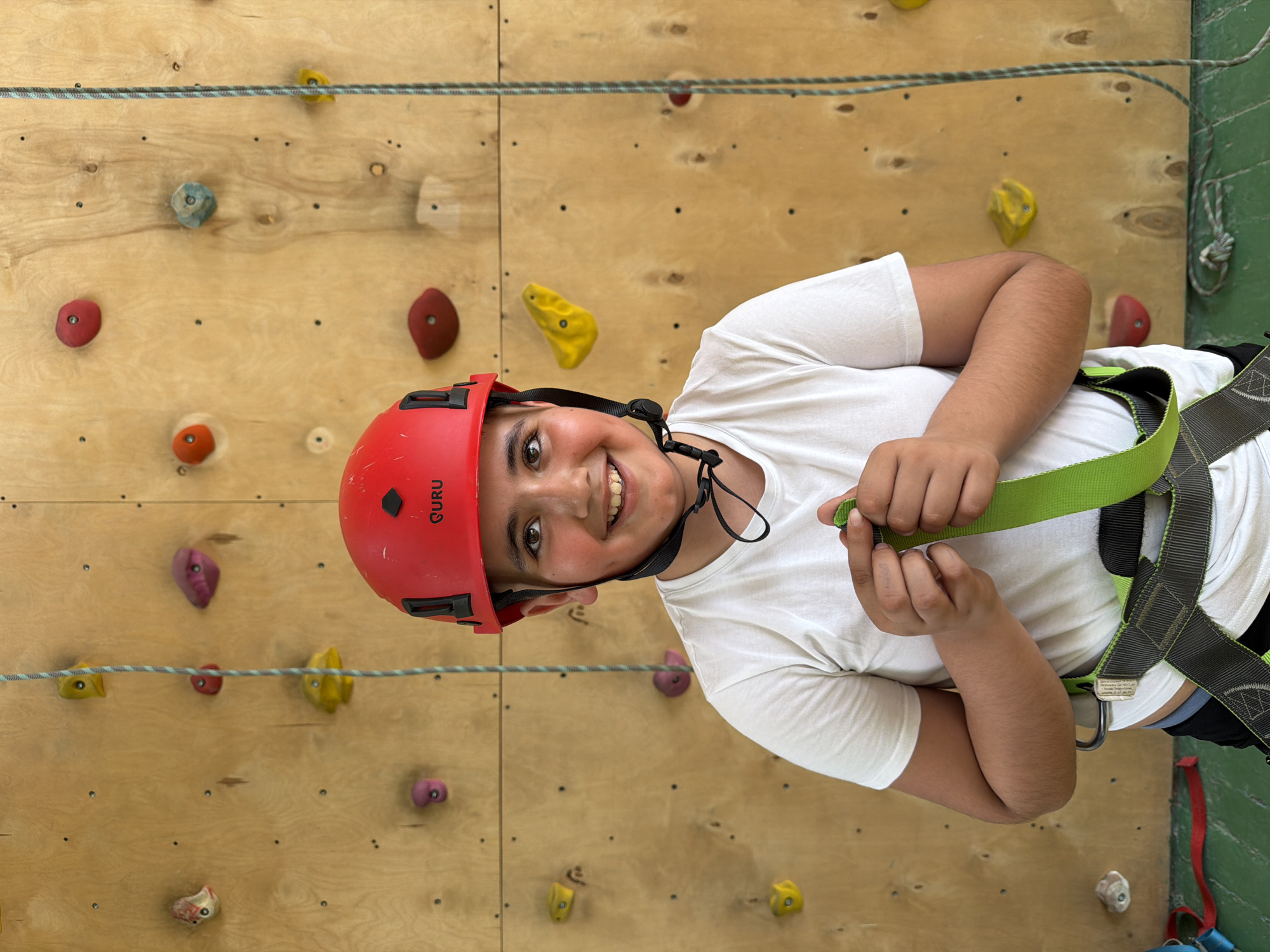 A young person wearing a red "GURU" branded climbing helmet and a white t-shirt, is suspended in climbing gear against a climbing wall with various colored holds. The climbing wall appears to be made of wood panels and has a rope strung across it. The location of the image is unknown, and I have no evidence to suggest it is in Izhevsk, Russia.