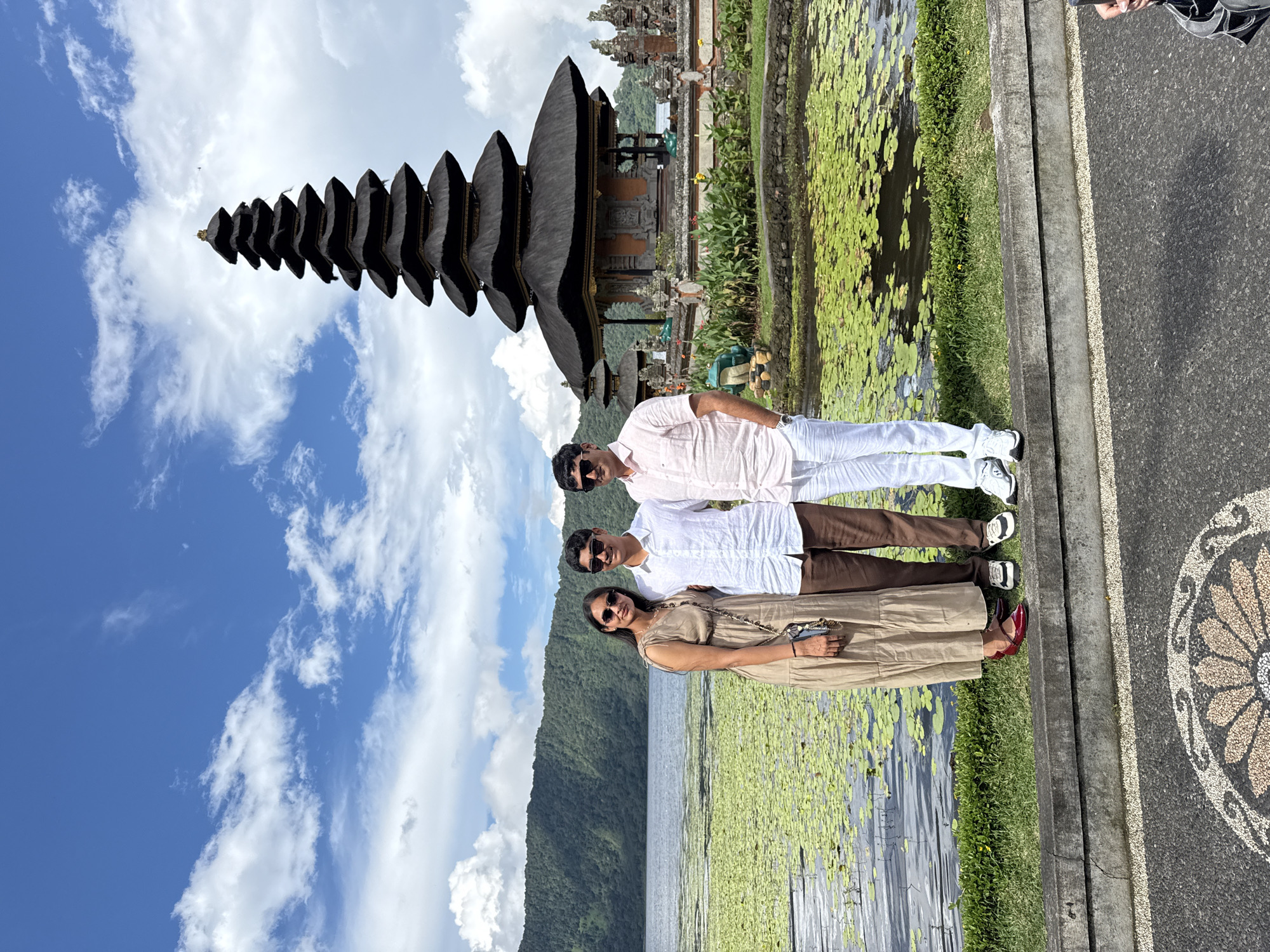 A medium shot depicts a family of three standing in front of the Ulun Danu Beratan Temple in Baturiti, Indonesia. The family is positioned on the right side of the frame, near the edge of a paved area. The woman in the front wears a tan dress and red shoes; the man behind her has a white shirt and brown pants; and the man in the back wears a pink shirt and white pants. They all wear sunglasses.
The Ulun Danu Beratan Temple, with its distinctive tiered roofs, is in the background to the left. In front of the temple is a lake covered with lily pads. A mountain range is visible behind the lake. The sky is blue with scattered clouds. A portion of a sidewalk with a circular design is visible to the right of the family.