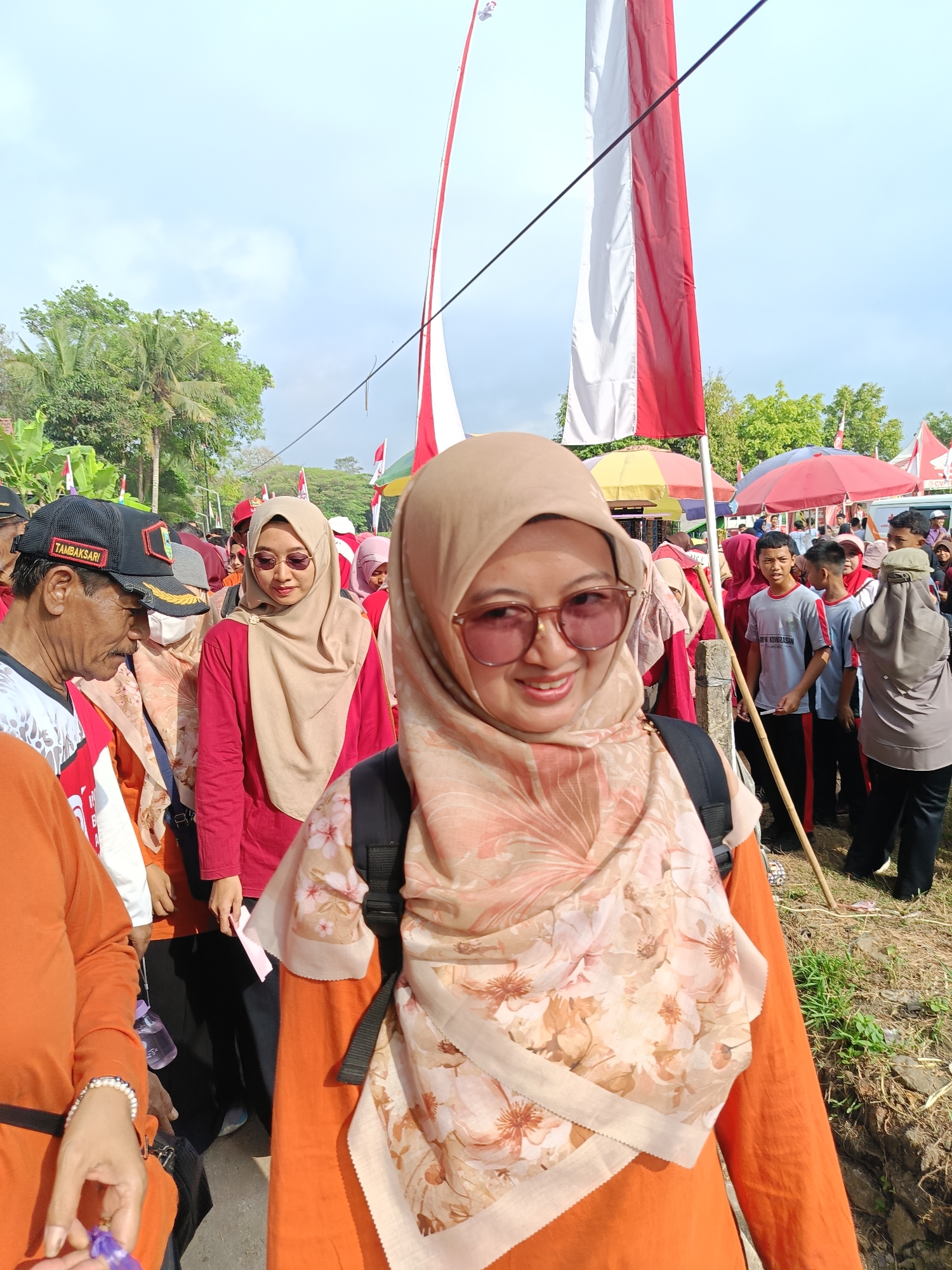 In the city of Mangli, Indonesia, a group of people are gathered outdoors, seemingly for a local event or celebration. The scene is filled with a festive atmosphere, indicated by the presence of Indonesian flags, a common sight during national holidays or community gatherings. In the foreground, a woman in a light brown hijab and an orange long-sleeved shirt is smiling at the camera. She is wearing sunglasses and a patterned scarf adorned with floral designs. Behind her, other individuals are visible, some wearing similar headscarves, suggesting a predominantly Muslim community.
To the left, a man with a mustache, wearing a black cap that reads "TAMBAKSARI", is looking downwards. He is dressed in an orange vest over a white shirt. In the background, a crowd of people are milling about, with some standing under colorful umbrellas, possibly for shade from the sun. The environment appears to be a dirt path or open area with some greenery and trees visible. The sky is bright and cloudy, suggesting daytime. The overall impression is a candid shot capturing a moment of community engagement in a pleasant, outdoor setting.