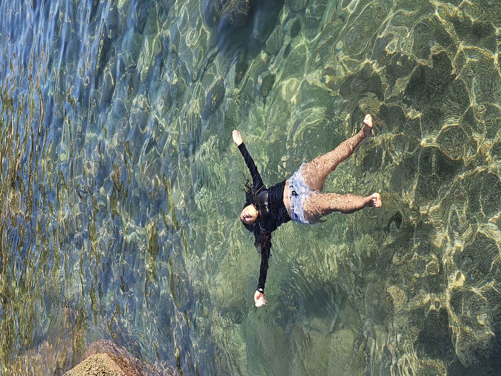 The image depicts a person floating in the clear, shallow, turquoise water of a beach located in Armação dos Búzios, Brazil. The person is lying on their back with their arms outstretched and their legs slightly apart. They are wearing a long-sleeved black top and denim shorts. The water reflects sunlight, creating shimmering patterns on the seabed. Some rocks are visible along the coastline. The image is taken from an elevated perspective, looking down at the person in the water.