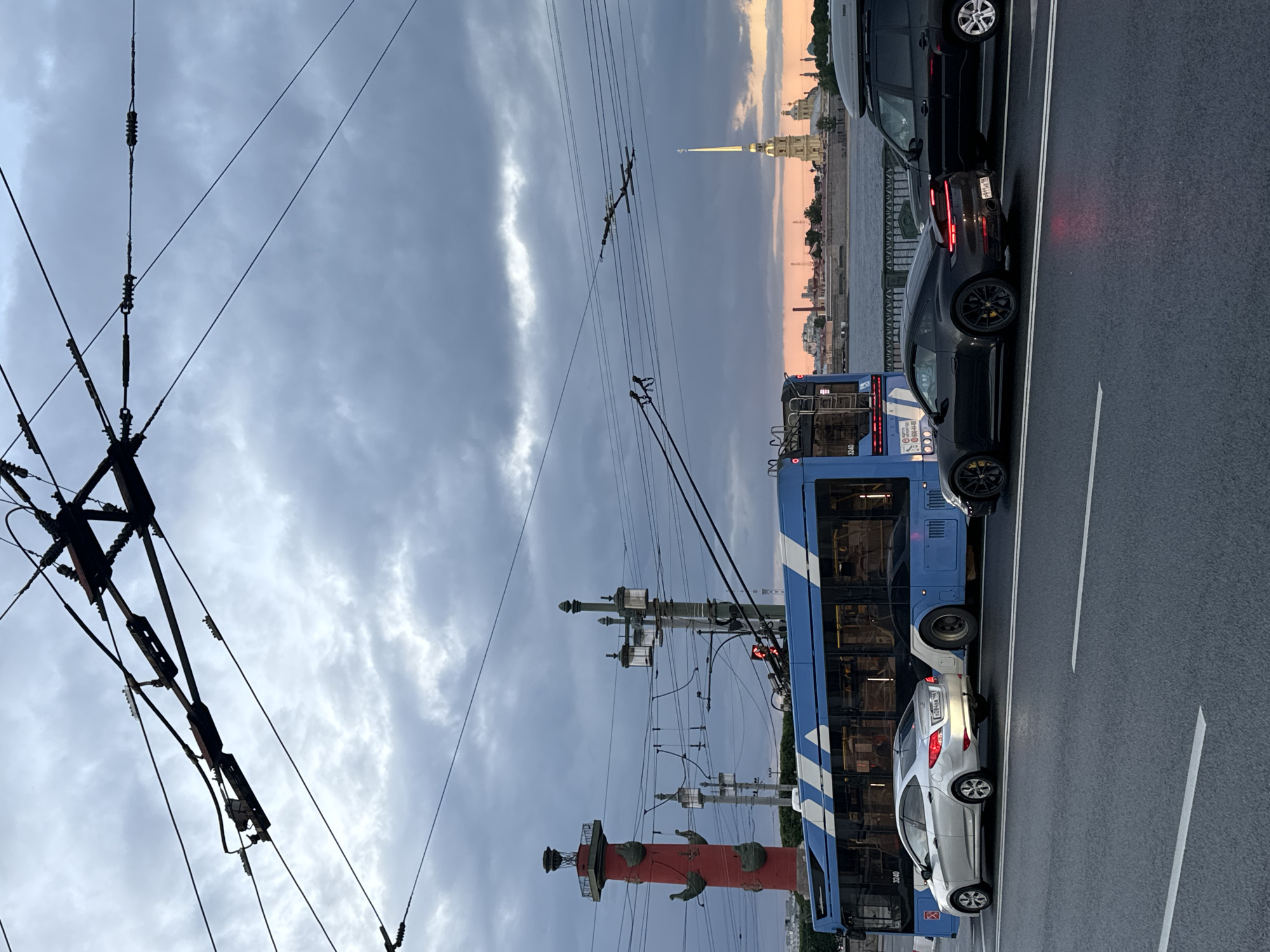 This image depicts a street scene in Saint Petersburg, Russia, with a view of the Peter and Paul Fortress in the background. The foreground features a road with vehicles, including a silver sedan, a black sports car, and a black SUV. A blue trolleybus is also visible. Overhead, intricate electrical wires for the trolleybus crisscross the frame, and a red Rostral Column stands on the left. The sky is overcast, with a hint of sunset colors near the horizon. The presence of the Rostral Column and the Peter and Paul Fortress strongly suggests the location is near the Neva River.