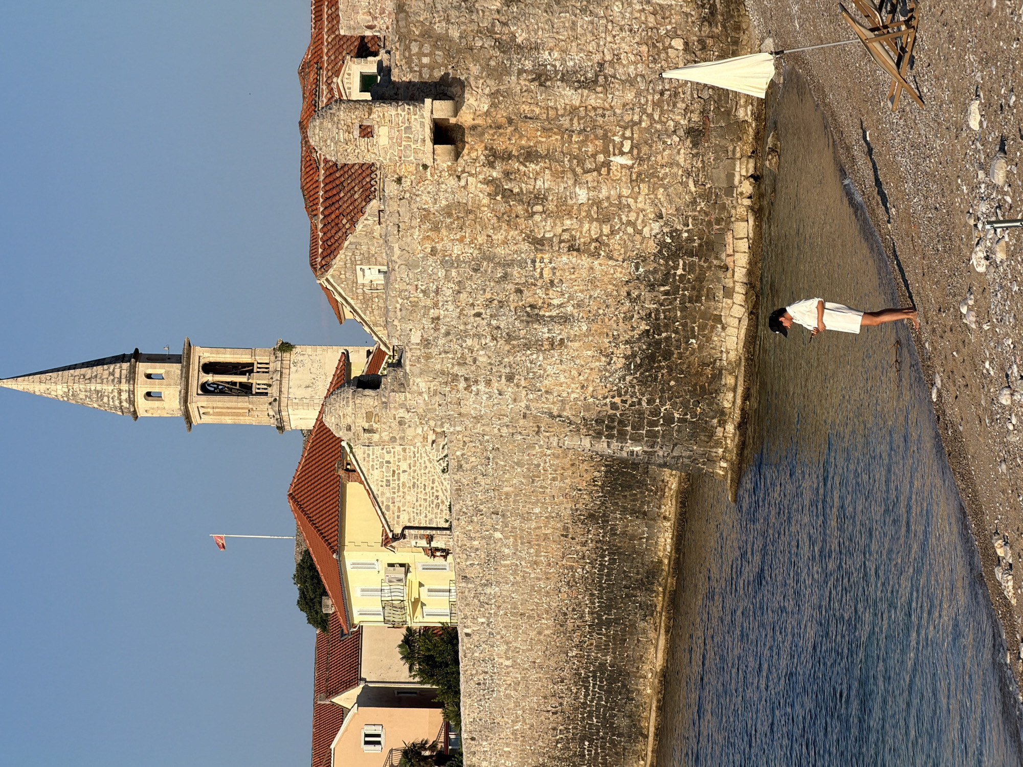 The image depicts the coastal city of Budva, Montenegro, specifically the old town walls and a section of the beach. In the background, the bell tower of the Church of St. John dominates the skyline, with a red flag flying from a flagpole to its left. Several buildings with red-tiled roofs are visible behind the stone wall. In the foreground, a woman wearing a white dress and a black baseball cap stands on the pebble beach near the water's edge, looking at her phone. Two wooden lounge chairs and a white umbrella are also present on the beach. The sea is calm and blue, with gentle waves lapping at the shore. The sun is casting long shadows, indicating it is likely late afternoon or early morning. The stone walls of the old town are aged and weathered, showcasing their historical significance.