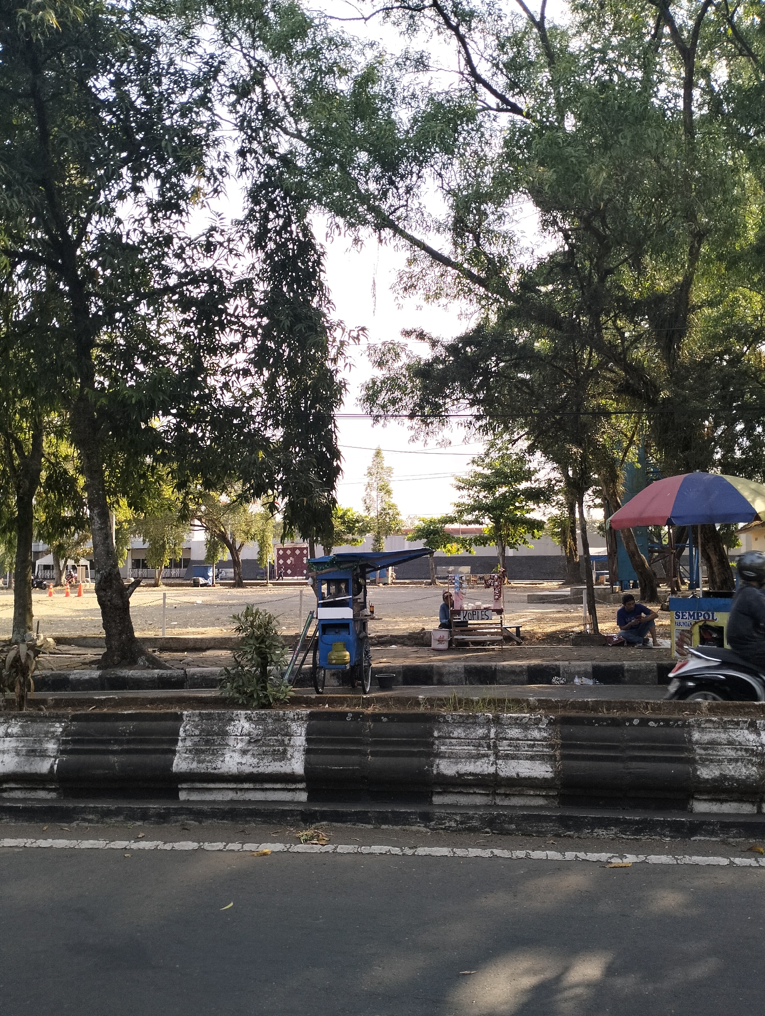 The image depicts an outdoor scene, likely a street or a park area, in Purbalingga, Indonesia. The foreground shows a paved road with a curb that has alternating black and white painted sections. Beyond the curb, there is a patch of dry grass and then a paved area.
Several trees with lush green foliage are prominent throughout the image, casting shadows on the ground. In the background, there are some buildings and structures, including what appears to be a long, low building with windows and a darker tiled roof. There are also some smaller stalls or carts set up, with one blue wheeled cart resembling a food or drink vendor.
People are visible in the scene. On the right side, a person is seated near a stall with a colorful umbrella. Further to the left, another person is seated near a stall with a sign that reads "KOPIES" (coffee). A motorcycle is passing by on the road in the foreground, with a rider wearing a helmet.
The overall impression is a moderately busy area with vendors and pedestrians, set against a backdrop of greenery and urban structures. The lighting suggests it might be late afternoon, with the sun casting long shadows.