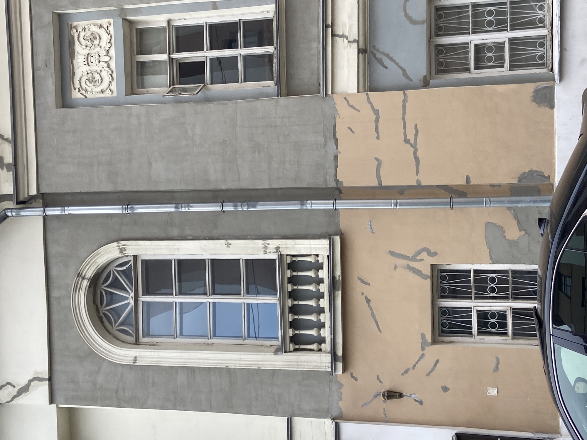The image shows the facade of a building in Moscow, Russia, with a mix of architectural styles and building conditions. The building is multi-story, and sections of the facade appear to have been recently renovated or repaired, with different colors of paint. There are several windows of varying shapes and designs. Some windows have ornate decorative elements. A metal drain pipe runs horizontally across the facade. A portion of a car is visible on the right side of the frame, reflecting the buildings across the street. The overall impression is one of an older building undergoing restoration or renovation.