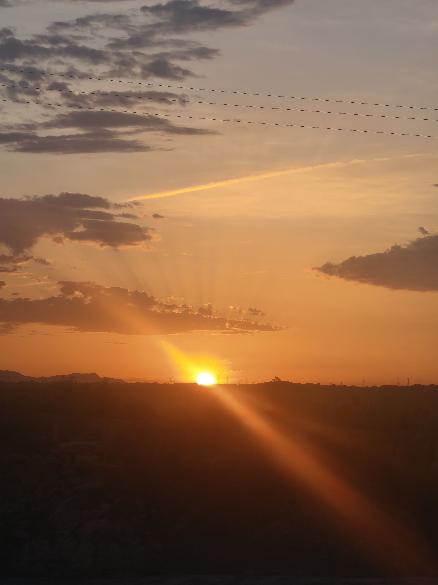 The image depicts a sunset over a rural landscape, likely on the outskirts of Riba-roja de Túria, Spain. The sun, a bright, intense orb, is setting near the horizon, casting long, golden rays across the sky. The sky is a gradient of warm oranges and yellows, transitioning to a softer peach and then a pale lavender towards the top. Wispy clouds, tinged with pink and gray, are scattered across the sky, with some larger, darker clouds present on the left side.
In the foreground, a dark, silhouetted landscape stretches across the bottom of the frame. This appears to be a sparsely vegetated area, possibly with some rocky terrain. The silhouette suggests rolling hills or a slight elevation. In the middle ground, a faint line of trees and structures can be discerned along the horizon, with what appear to be power lines stretching across the upper portion of the sky. There are no people or animals visible in the image. The overall impression is one of a tranquil, natural scene at the end of the day.