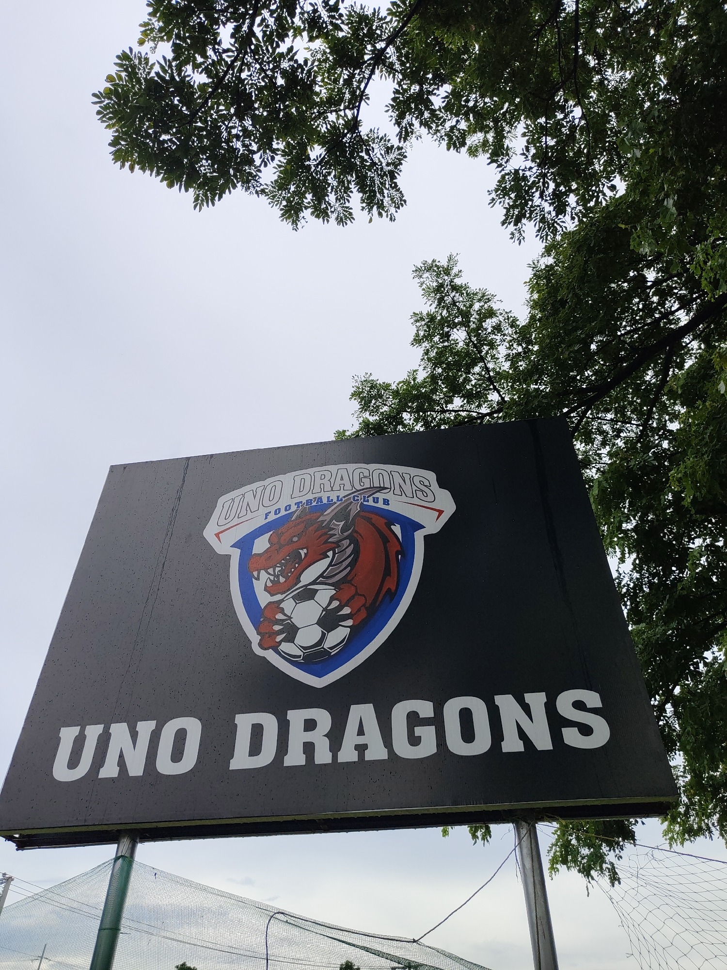 The image displays a sign for "UNO DRAGONS FOOTBALL CLUB" located in Davao City, Philippines. The sign features a red dragon emblem with a soccer ball, set against a dark background. The sign is tilted upwards, with green foliage from trees visible at the top and right of the frame. Below the sign, a chain-link fence or net is partially visible, suggesting a sports field or court. The sky is overcast.