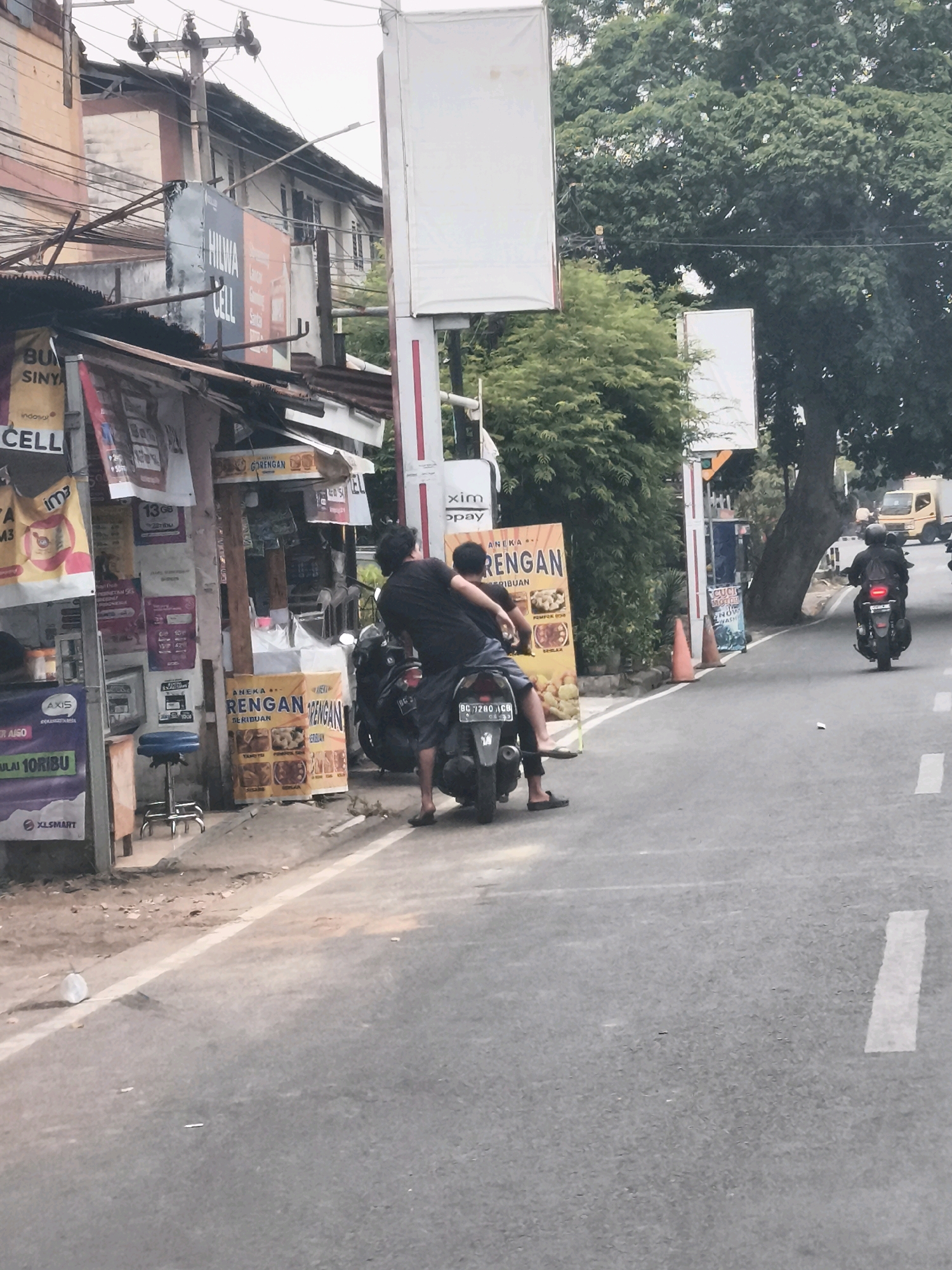 The image depicts a street scene in Palembang, Indonesia, likely during the daytime under overcast skies. In the foreground, two individuals, both appearing to be male, are seated on a parked motorcycle. The person in the back is leaning forward, embracing the person in the front. Both are dressed casually. The motorcycle has a license plate that reads "BG 7280 ICG".
To the left of the motorcycle is a small roadside stall or shop. The stall has a corrugated metal roof supported by wooden poles. Various posters and signs are attached to the front of the stall, some with text in Indonesian, including what appears to be advertisements for phone services and food. A blue stool is visible near the stall.
The street itself is paved with asphalt, with a white line marking the edge of the road. Further down the street, another motorcycle with a rider is visible, moving away from the viewer. A yellow truck is also present further in the distance on the right side of the road. Tall trees with dense green foliage line the right side of the street, providing shade. Several utility poles with wires crisscross above the buildings. The general environment suggests an urban or semi-urban setting.
