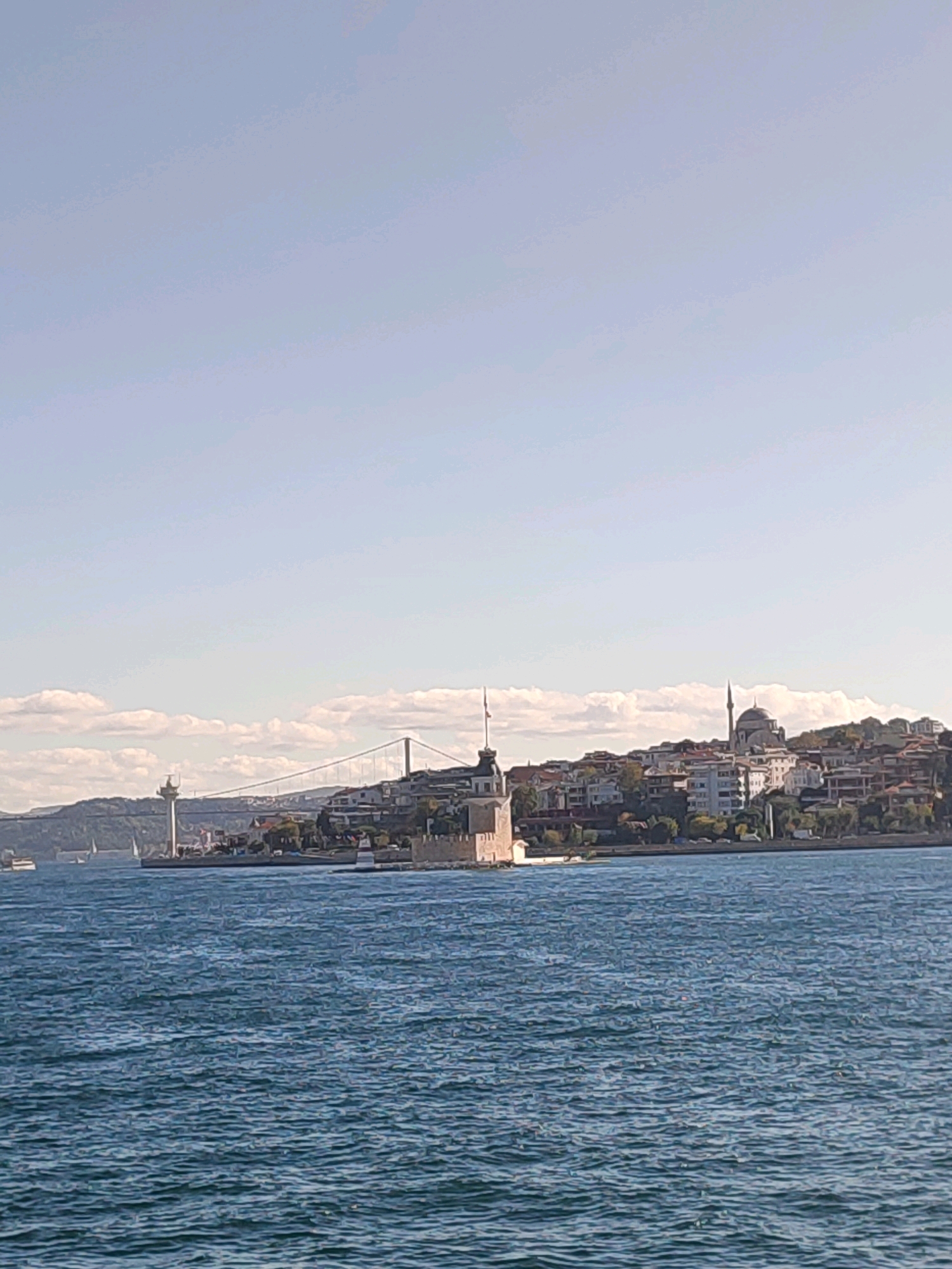 The image shows a panoramic view of the Üsküdar coastline in Istanbul, Turkey, from the water. In the foreground, the blue water of the Bosphorus is rippled by a gentle breeze. In the middle ground, on the shore, stands the Maiden's Tower, a historic structure with a cylindrical base and a tower on top, flying a flag. To the right of the Maiden's Tower, a prominent mosque with a dome and a minaret is visible, its architectural style indicative of Ottoman influence. Behind the Maiden's Tower and the mosque, a dense collection of buildings, typical of a city, climbs up a hill, interspersed with trees. Further in the background, a suspension bridge spans the water, connecting the Üsküdar side to the other side of Istanbul. The sky above is a clear blue, with a few scattered white clouds. The overall scene depicts a sunny day, highlighting the picturesque cityscape of Üsküdar.