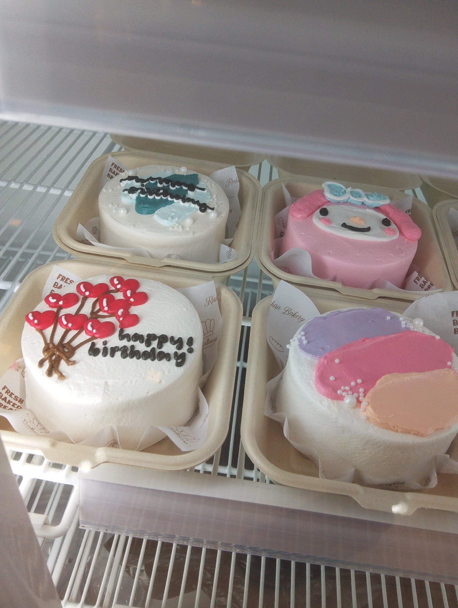 Four small cakes are displayed in individual light brown cardboard containers, placed on the white wire shelves of a refrigerator. The cakes are decorated with frosting and edible embellishments.
The cake in the front left is white, with a bouquet of red heart-shaped balloons made of frosting, tied with brown string. The words "happy! birthday" are written in black frosting below the balloons.
To the right of that cake is a pink cake decorated to resemble a character, likely from a cartoon, with large ears, a bow on its head, and facial features.
The cake in the back left is white with blue and black frosting decorations resembling water and waves, and white pearl-like candies.
The cake in the front right is white with three large, colorful frosting shapes on top: purple, pink, and pale orange. Small white pearl-like candies are scattered on the frosting.
Small white paper liners with printed text and logos are visible within the cardboard containers. The environment appears to be the interior of a refrigerated display case or storage unit, with visible white metal shelving. The lighting is bright and diffused.
There is no visible indication of the city of Manaruwi, Indonesia, within the image.