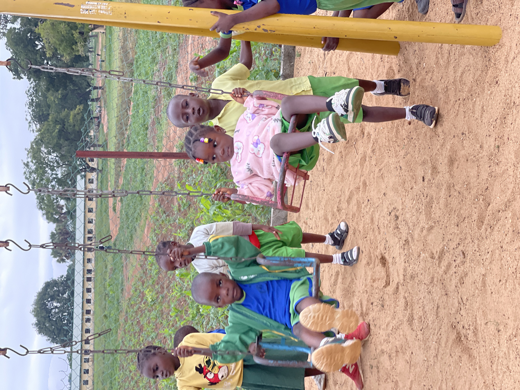 The image depicts a vibrant outdoor scene of several young children playing on a swing set in Akwanga, Nigeria. The setting is a playground with sandy ground beneath the swings and a chain-link fence separating the play area from a grassy field and trees in the background. A long, light-colored building with multiple windows is visible in the distance, suggesting a school or community facility. The sky is overcast, indicating a cloudy daytime.
At the center, a young girl with braided hair adorned with colorful beads, wearing a light pink, long-sleeved top with bunny motifs, sits on a swing and looks directly at the camera. Behind her, a boy in a yellow short-sleeved shirt and green shorts stands, holding the swing chains, also with his gaze towards the camera. To their right, another boy in a green jacket over a blue shirt and green shorts is seated on a swing, sporting light-colored sneakers, and looking at the viewer. Other children are partially visible around the swing set, some on swings and others standing near the structure. One child on the far left wears a yellow t-shirt with a cartoon character print.
The swing set itself is made of yellow metal, with chains supporting the seats. Prominently displayed on the main yellow crossbeam are white text markings: "MUNICIPAL INTERVENTION," "YEAR 2013," and "SDG AD AKWANGA." This text indicates the swing set was part of a municipal project related to Sustainable Development Goals, installed in Akwanga in 2013. The children appear calm and engaged in their play, with some exhibiting curious expressions as they look towards the camera.
