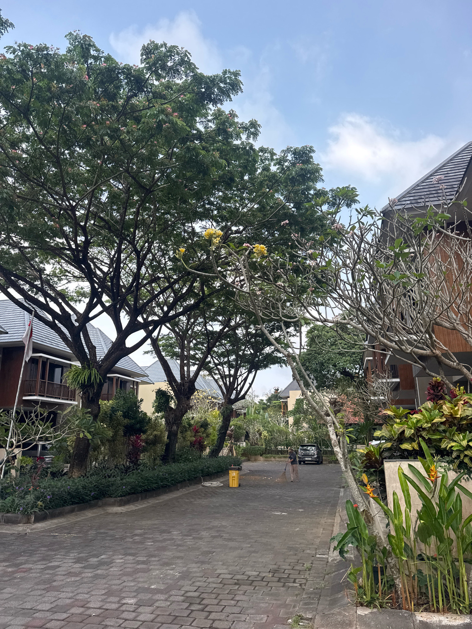 The image depicts a paved street lined with buildings and lush greenery in Kuta Selatan, Indonesia. The buildings appear to be modern, with wooden accents and tiled roofs. Several mature trees with dense foliage and some blooming flowers are scattered throughout the scene, casting shadows on the street. In the middle ground, a person is sweeping the street with a broom, while a car is parked nearby. The sky is partly cloudy, with a mix of blue sky and white clouds. The overall impression is one of a well-maintained residential or resort area.