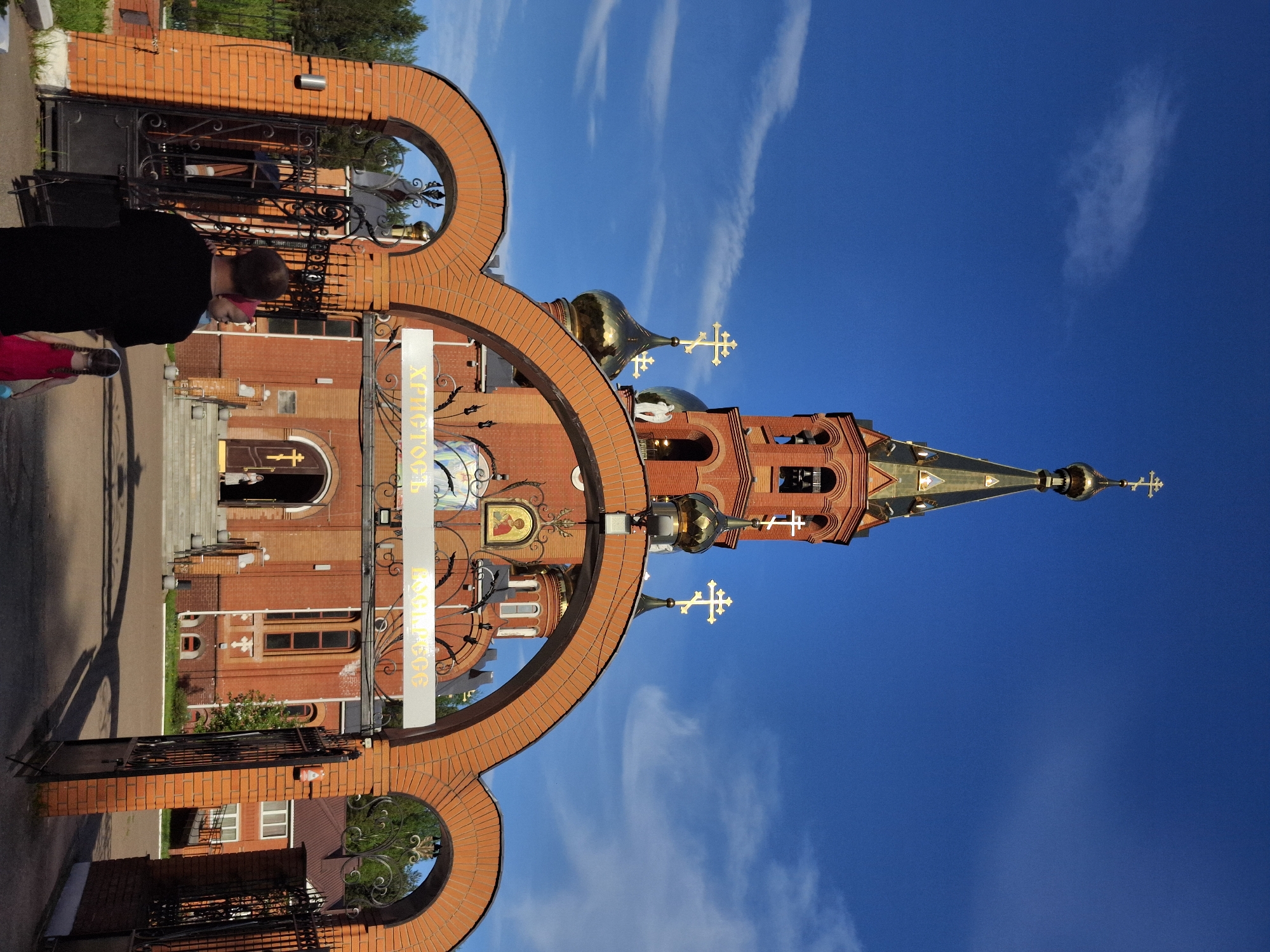 A vertical outdoor shot shows the Church of the Resurrection of Christ located in the city of Chaikovsky, Russia. The church, constructed of red brick, features golden domes with crosses atop. The church is framed by a large brick archway with decorative metalwork. In the foreground, a woman in a black shirt and a child in pink clothing are standing on the pathway leading to the church. The sky above is a clear blue with wispy white clouds. An open iron gate is at the left foreground.