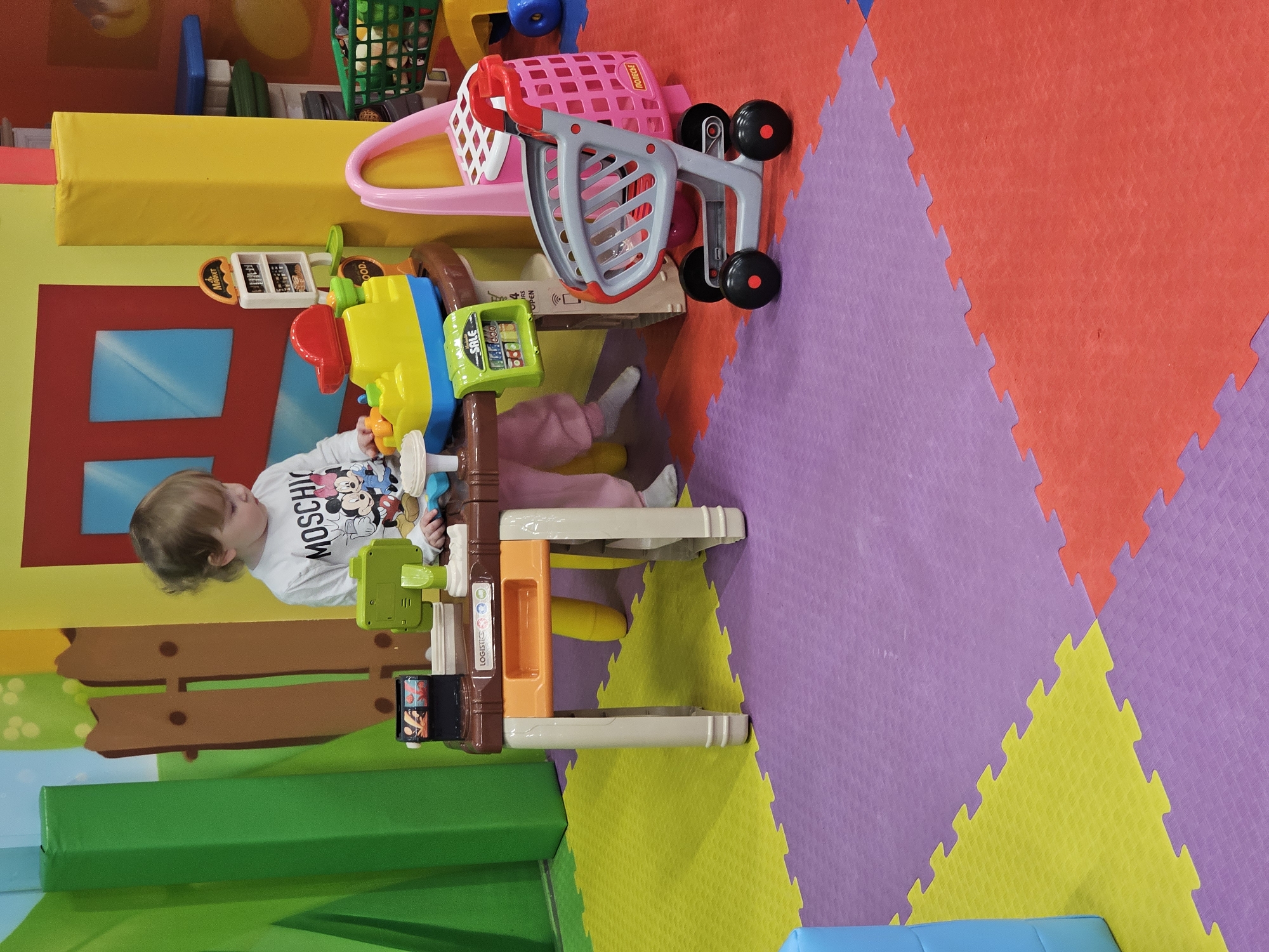 A young child wearing a white Moschino shirt and pink pants is playing at a toy checkout counter. The counter is colorful with various toy elements, including a scanner and buttons. Nearby is a toy shopping cart. The scene takes place on a floor covered with interlocking foam tiles in purple, red, yellow, and other colors. The location is Khimki, Russia.