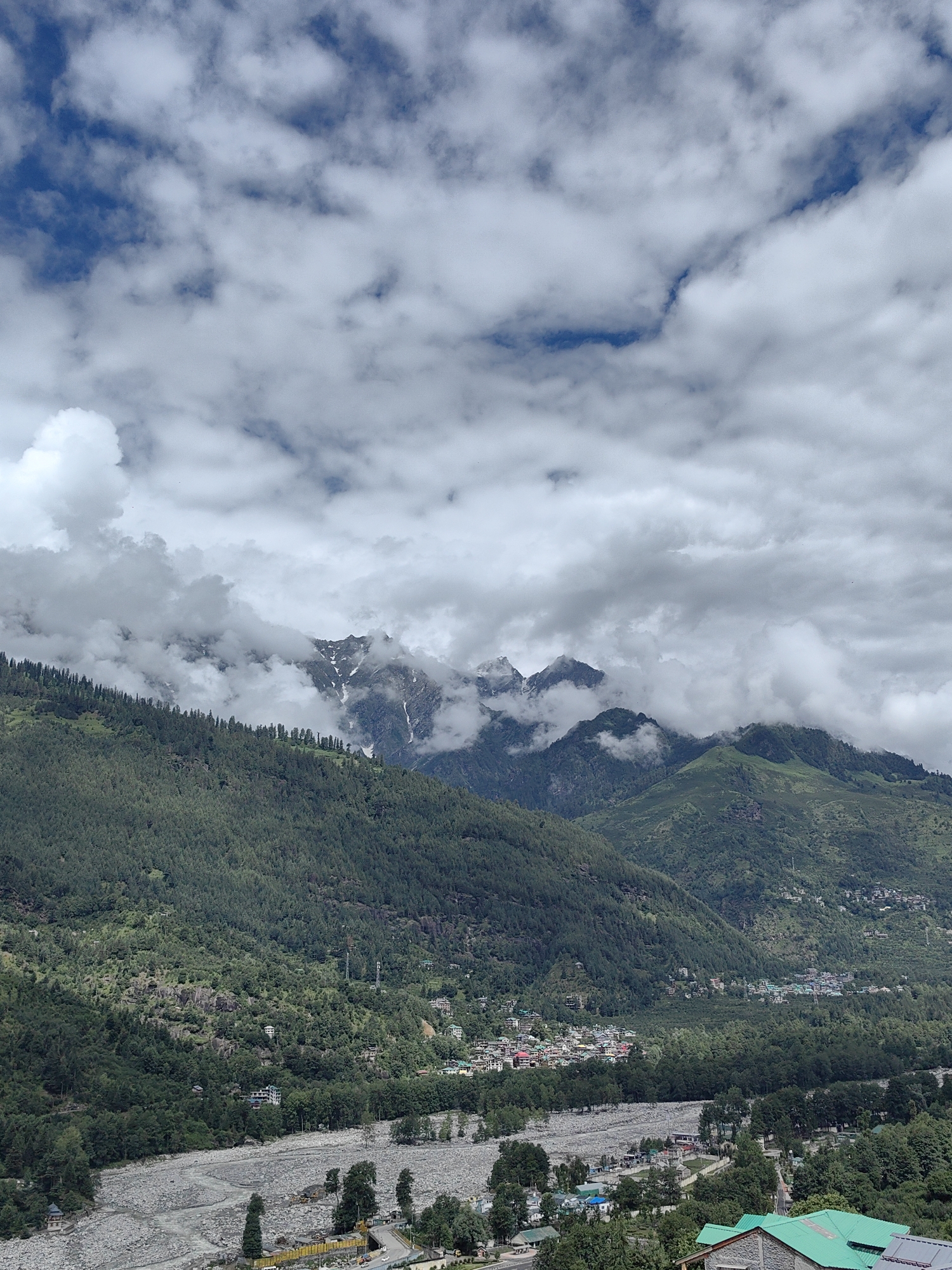 The image depicts a mountainous landscape with a valley and a river, likely in the vicinity of Vashisht, India. The sky is filled with dramatic, fluffy white clouds against a backdrop of blue. In the midground, steep, heavily forested mountains rise, with some peaks revealing snow-capped summits peeking through the clouds. A small town or village is nestled within the valley, characterized by clusters of buildings with colorful roofs. A wide, rocky riverbed, with a winding stream of water, flows through the valley floor. The foreground shows more of the valley floor with some trees and buildings, including one with a green roof. The overall impression is of a serene, natural environment with a human settlement integrated into the landscape.