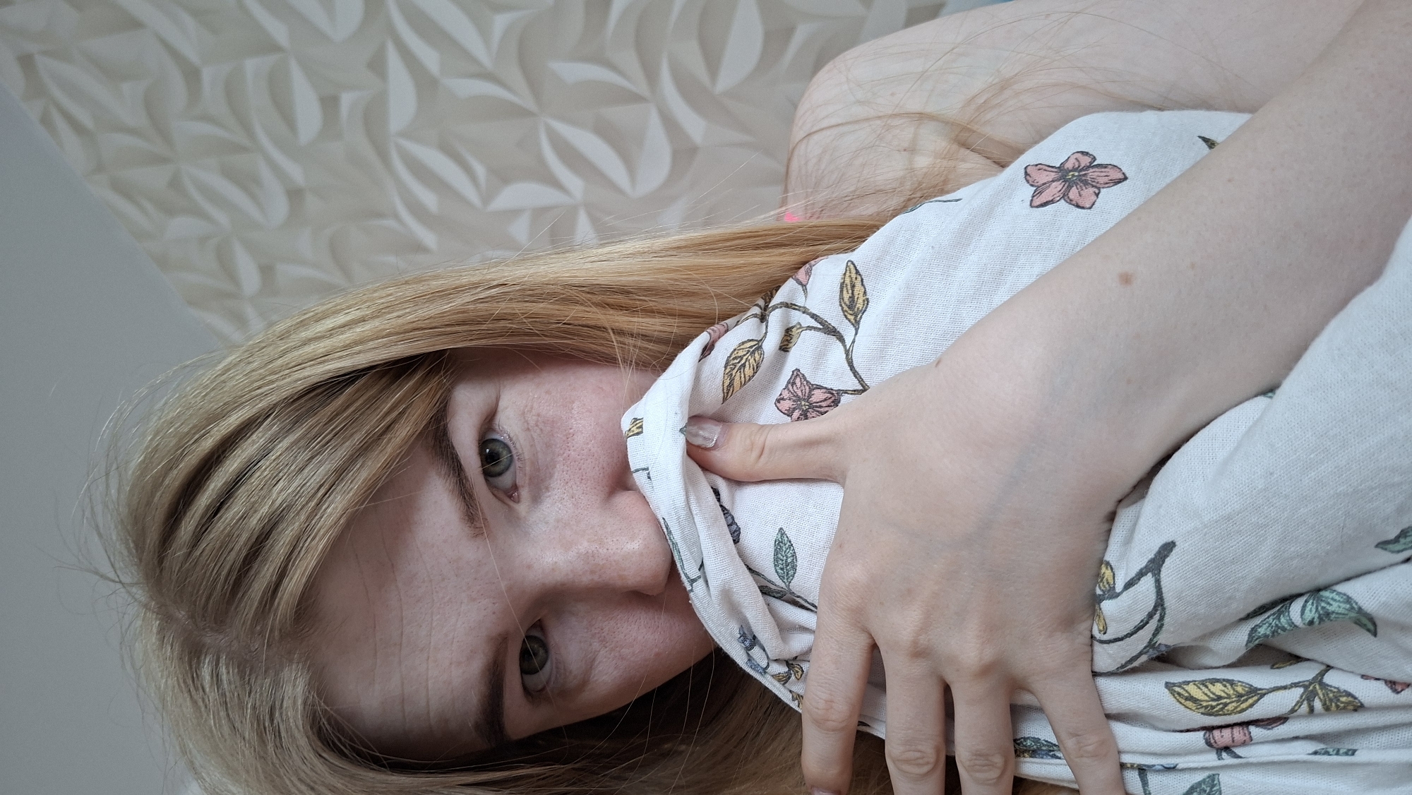 A woman with fair skin and light brown hair is lying on her side, holding a white patterned cloth to her face. The cloth features floral designs in various colors. Her eyes are open and she is looking directly at the camera. Part of a pink object is visible at the top of her head, possibly related to her hair. The background consists of a wall with a geometric pattern at the top and a plain painted wall at the bottom. The image is rotated counterclockwise. The location is in the city of Chaikovsky, Russia.