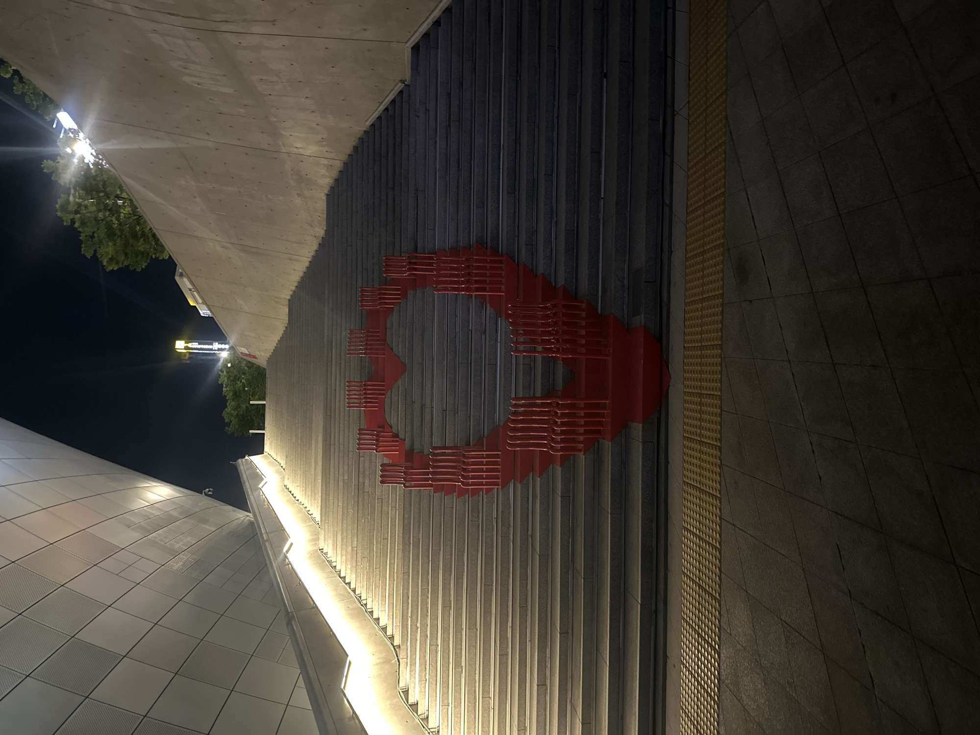 The image depicts a tilted upward view of an exterior urban scene at night, likely in Seoul, South Korea. The primary focus is a tall, vertical gray wall with a large, red, stylized geometric shape resembling a heart with an opening. The heart is formed from multiple horizontal red bars arranged in layers. The wall is composed of vertical concrete or stone ribs, partially illuminated by artificial lighting along its base. To the left, a curved white building with a tiled surface can be seen. To the right of the wall is a textured yellow tactile paving strip along the edge of a dark, paved ground surface. The sky is mostly dark, with some trees and distant buildings visible in the upper left corner, illuminated by streetlights. There are no people visible in the frame.
