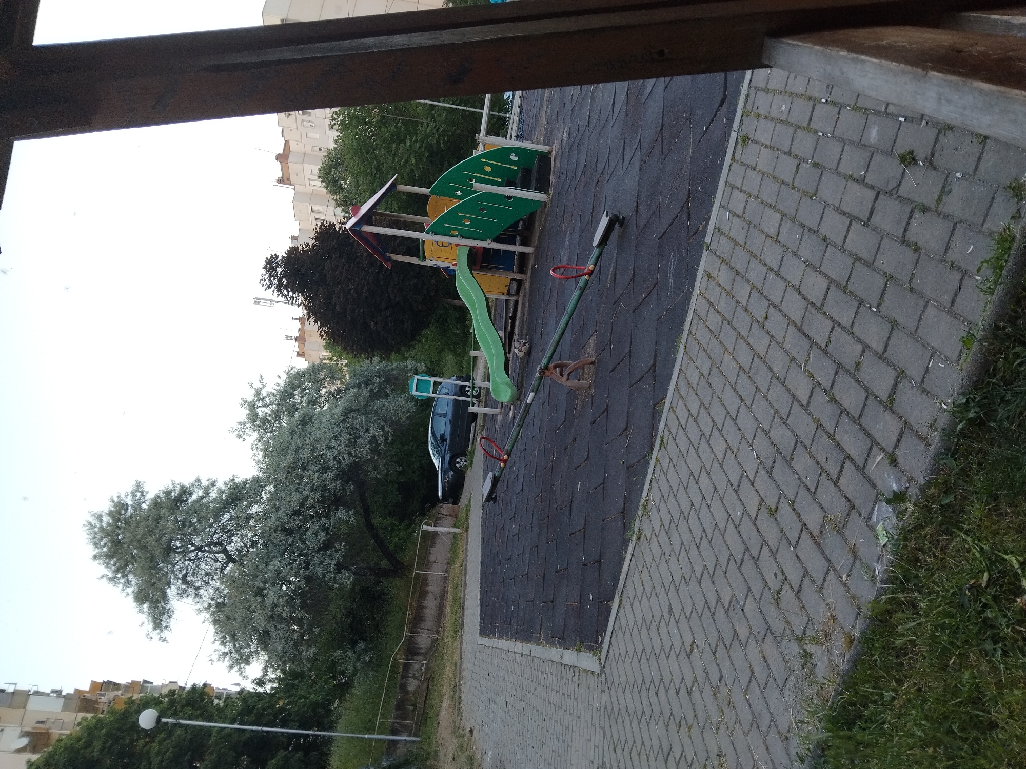 The image depicts a playground located in Stara Zagora, Bulgaria. The playground features a slide and seesaw. A car is parked nearby. The ground is covered in gray paving stones and black rubber tiles. Buildings and trees are visible in the background. The image is taken from a partially obstructed perspective, with a wooden structure in the foreground.