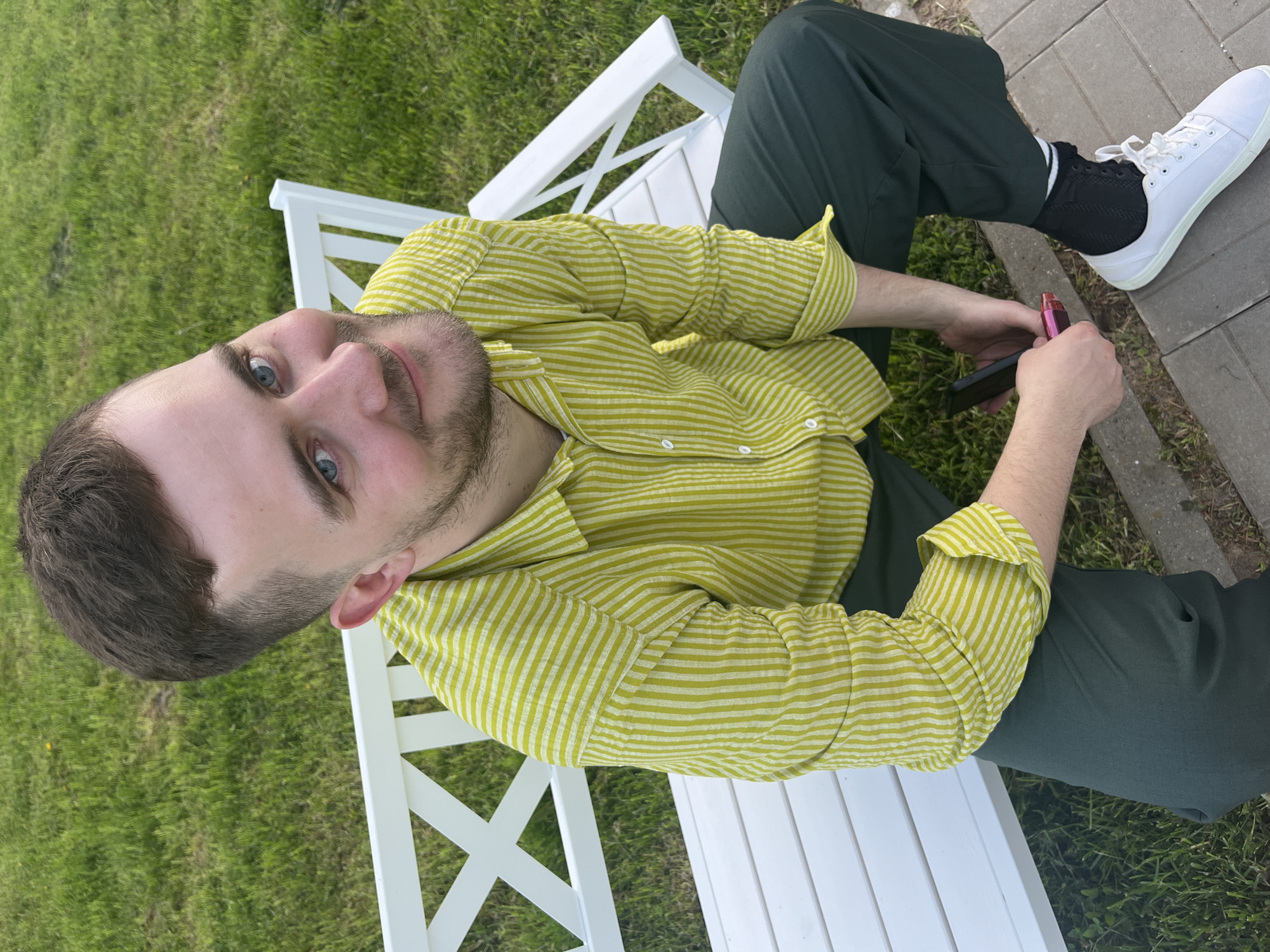 A young man with short brown hair and a beard sits on a white wooden bench, holding a black smartphone and a pink vaping device. He is wearing a light green striped button-down shirt and dark green pants. His left foot, clad in a white sneaker and black sock, is visible. The bench is located on a grassy area adjacent to a paved walkway. This is presumably taken in the city of Алопово, Russia.