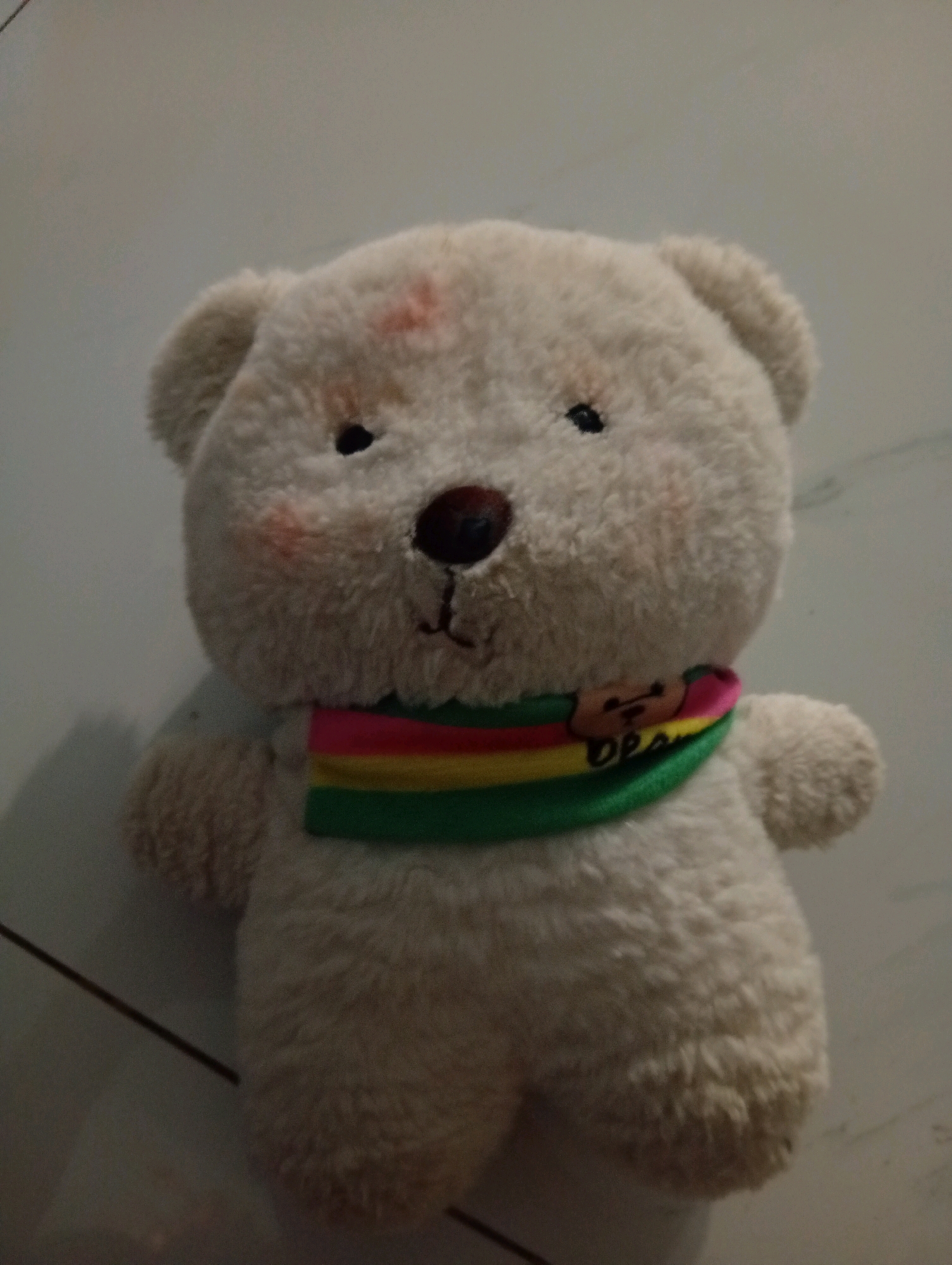 A fluffy, cream-colored teddy bear is positioned on a grey, tiled floor. The bear is facing forward and has a small, dark brown nose and two black eyes. There are faint pink marks on its cheeks and forehead. It is wearing a scarf with horizontal stripes in green, yellow, red, and pink. The scarf has a small, brown wooden charm with a teddy bear face on it and the word "Design" written below. The lighting in the scene is dim, creating shadows on the floor to the left of the bear. The overall environment appears to be indoors, likely a residential setting in Samarinda, Indonesia.