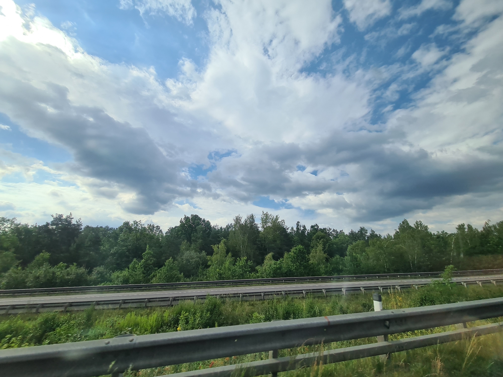 The image depicts a view of a highway bordered by a dense green forest under a cloudy sky. The highway has two lanes in each direction, separated by a metal guardrail. The foreground shows another guardrail, suggesting the photo was taken from a vehicle. The forest is lush and green, with a variety of trees. The sky is filled with a mix of white and grey clouds, with some patches of blue sky visible. The scene appears to be a daytime shot, possibly taken in the late spring or summer. There are no people or animals visible in the image. The location is identified as Loipersbach, Austria.
