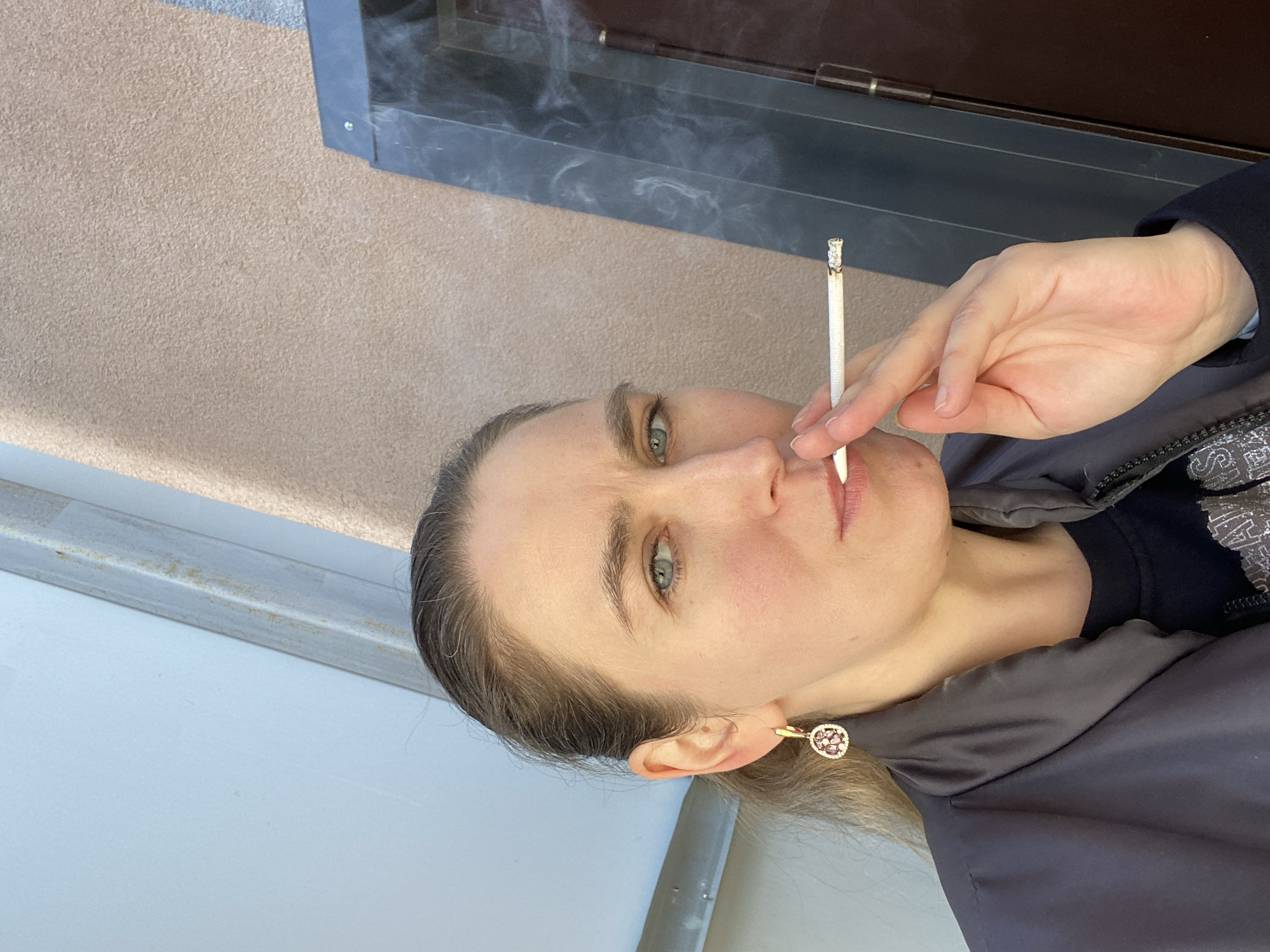 A woman with light skin and blue eyes is holding a lit cigarette to her lips. She is wearing a dark jacket and a gold earring with a decorative stone. She is outside in Yekaterinburg, Russia, and the background shows a building exterior with a window and a textured wall.