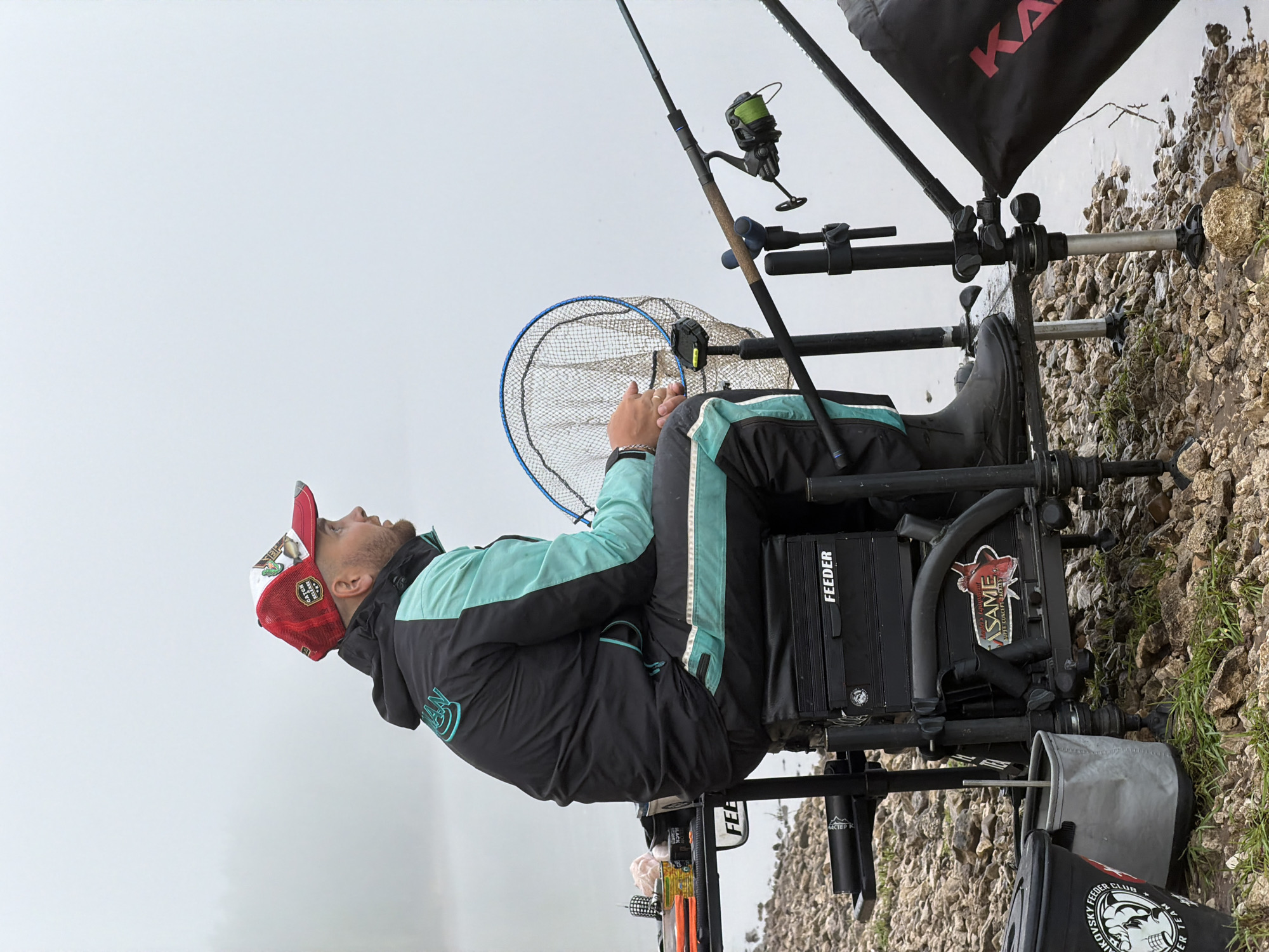 The image, rotated 90 degrees counter-clockwise, captures a man engaged in feeder fishing by a body of water, likely in Noviy, Russia. The scene is set on a rocky and pebbly bank with sparse patches of grass, bordering calm water that reflects the uniformly light, overcast, or foggy sky above. The diffused lighting suggests either early morning, late afternoon, or simply a heavily clouded day.
The primary subject is a man, seen from the waist up, seated comfortably on a specialized black fishing chair or box. He is dressed in functional fishing attire, including a black and teal waterproof jacket with reflective stripes and black trousers. A prominent red and white mesh cap adorns his head, featuring fish graphics and the texts "CATCH OR DIE" and "FISHING TEAM." His expression is one of calm concentration as he looks upwards or out towards the water, hands clasped, appearing to be patiently waiting for a bite.
Around the man is his fishing setup. A fishing rod, equipped with a reel spooled with bright green line, rests on a rod stand. Another fishing rod is visible nearby. A blue-rimmed landing net is positioned within easy reach. The robust fishing chair/box serves as his base, holding various attachments and supports for the rods and other gear. Visible branding on the equipment includes "FEEDER" printed on the chair and other items, "ASAME" accompanied by a fish logo, and "MACTEP K" (likely "Master K"). A black bag or bucket near the chair displays "FEEDER FISHING CLUB" with a skull emblem, and another partially visible black bag in the background shows "KA". The overall impression is one of dedicated and well-equipped angling.
