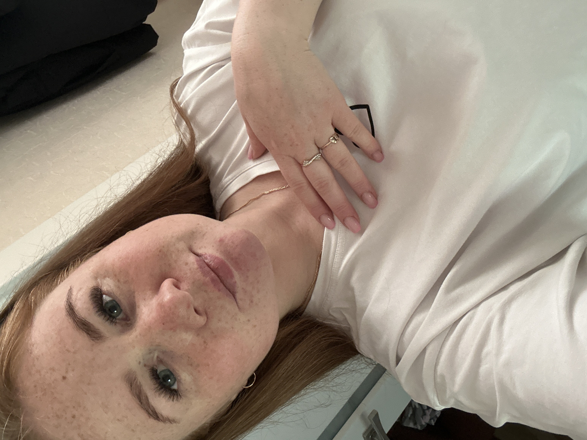 A photograph depicts a woman with fair skin and freckles, lying down and facing the camera. She has green eyes and reddish-brown hair. She is wearing a white t-shirt. She has rings on her fingers and a necklace around her neck. The background includes a light-colored surface and a dark-colored object. The location is not specified in the image. The image does not explicitly connect to Chaikovsky, Russia.