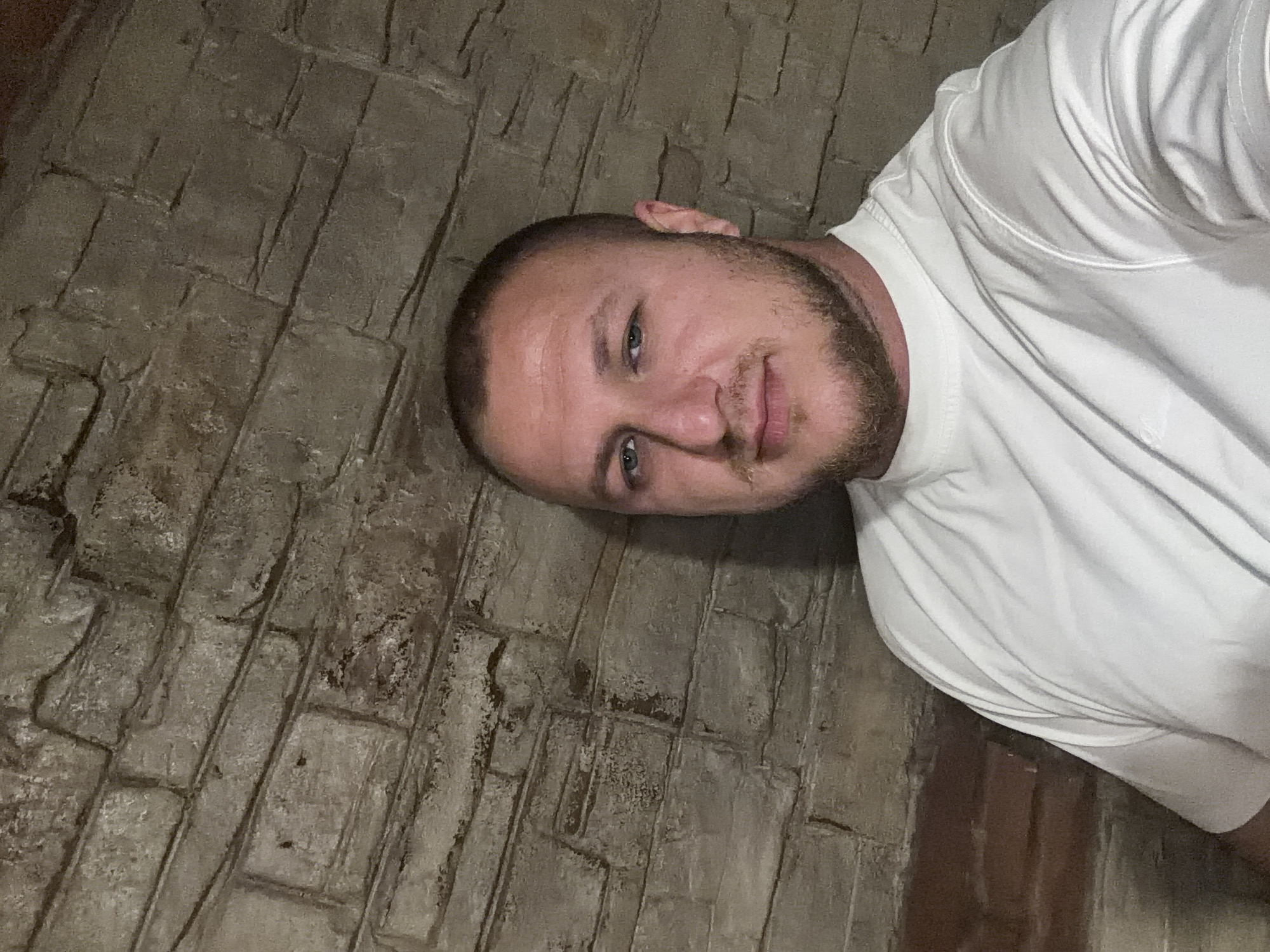 The image features a man taking a selfie against a textured, stone or brick-like wall in Asenovgrad, Bulgaria. The man has short, light-brown hair and a trimmed beard, with light-colored eyes. He wears a plain white short-sleeved t-shirt, on the right sleeve of which a faint "adidas" logo is visible. He looks directly at the camera with a neutral to slightly serious expression.
The background wall appears to be made of irregularly shaped, dark grey and brown blocks or stones, possibly part of an older building or an interior with a rustic finish. The mortar lines are clearly visible, highlighting the rough texture of the surface. The lighting is dim and somewhat uneven, casting shadows and suggesting an indoor setting or low-light conditions outdoors. No other distinct objects or activities are present. The overall atmosphere is muted due to the dim lighting and the dark tones of the wall.