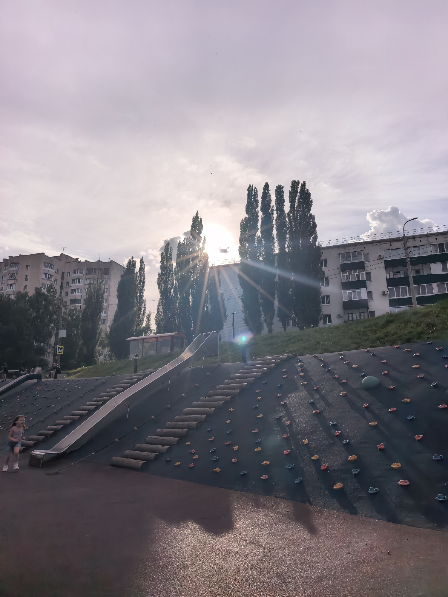 This is an eye-level, full shot of an outdoor playground on a cloudy day in Sterlitamak, Russia. The sun is peeking through the clouds, creating a bright glare in the center of the frame and casting long shadows across the playground. The playground features a slide, a climbing wall with colorful grips, and a series of wooden log steps. A young girl in a pink top and blue shorts is running on the lower part of the playground. In the background, there are tall, slender trees and multi-story apartment buildings, indicating an urban setting. The sky is a soft purple and grey, with a few scattered birds visible. The overall atmosphere is bright and slightly hazy, suggesting it is either early morning or late afternoon. There is no visible text in the image.
