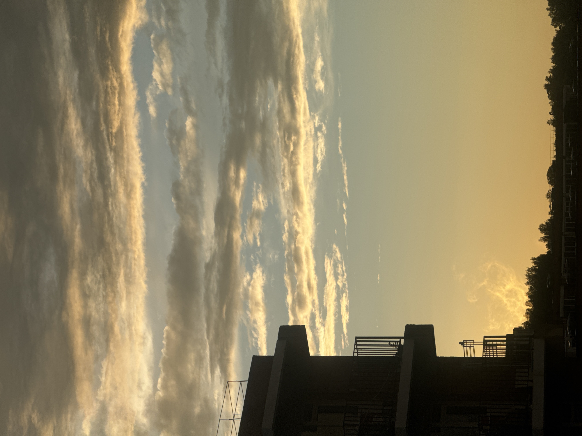 The image depicts a silhouetted building with balconies against a sky filled with dramatic clouds during a sunset. The building, appearing to be a multi-story structure, is dark and its architectural features, including railings and balconies, are clearly defined against the lighter background. The clouds are layered, with streaks of golden light illuminating them from below, suggesting a low sun angle. The lower portion of the image shows a line of trees and what appear to be other buildings, all silhouetted against the twilight sky. The overall scene is one of atmospheric beauty at dusk. The location in the city of Маркова, Russia is suggested by the presence of what could be typical Russian residential or commercial architecture and the characteristic northern sky.