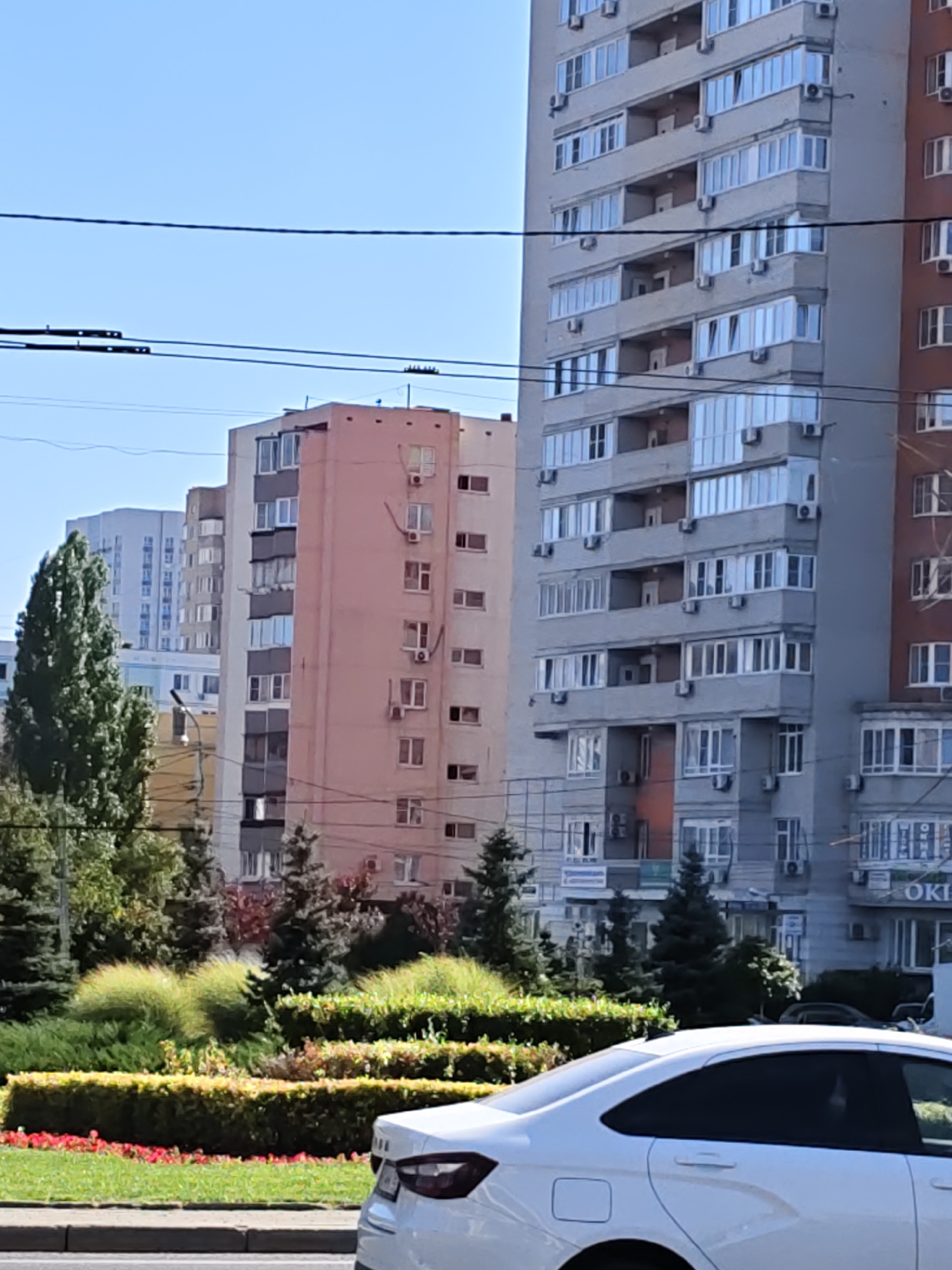 The image shows a cityscape with several apartment buildings of varying heights and architectural styles. In the foreground, the rear of a white sedan is visible, parked on the side of a road. Behind the car, a landscaped area with bushes, trees, and red flowers is present, indicating a well-maintained urban green space. The buildings are characteristic of Soviet-era or post-Soviet architecture, with multiple floors, balconies, and prominent air conditioning units. One building is painted a light pink, while others are grey or brown. In the background, a taller, modern-looking apartment building with a lighter facade is visible. The sky is clear and blue, suggesting a sunny day. The overall scene depicts a typical urban environment, likely in a city with a temperate climate. The presence of air conditioning units suggests a region that experiences warm summers. The text "OKI" is visible on a sign on one of the buildings, which could potentially be a business or a building name. The composition and subject matter do not provide definitive clues to pinpoint a specific location within Rostov-on-Don without further information.
