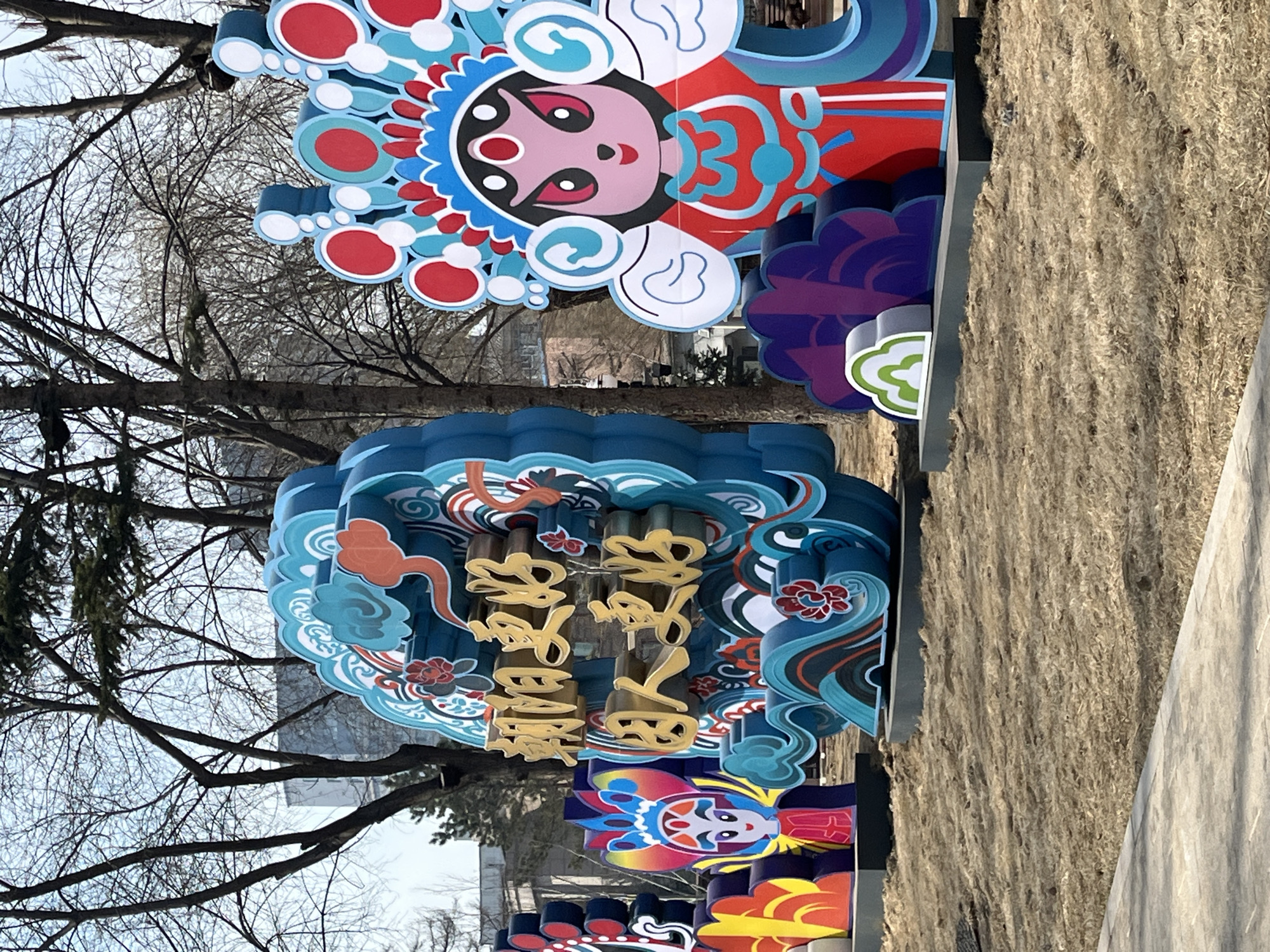 This outdoor shot, taken in Changchun City, China, captures a display of Chinese cultural ornamentation near bare trees. Dominating the view is a large, vertically oriented art installation featuring a depiction of a figure from Chinese opera. The figure has a stylized face with red and pink accents, a detailed headdress with various colors including red, white, and blue, and is set against layers of decorative shapes. Below the figure, there are Chinese characters rendered in a golden hue, arranged in vertical columns, possibly indicating the name of a place or a celebratory greeting for the New Year.
The installation is surrounded by stylized blue waves and cloud-like motifs, reinforcing the festive nature of the decoration. The barren branches of nearby trees create a stark contrast to the colorful display, indicating it might be winter or early spring. A wall, textured with moss or fiber, is seen on the right side of the frame, setting off the vibrant colors of the installation. The photograph appears to have been taken at ground level, with a slight upward angle towards the artwork.